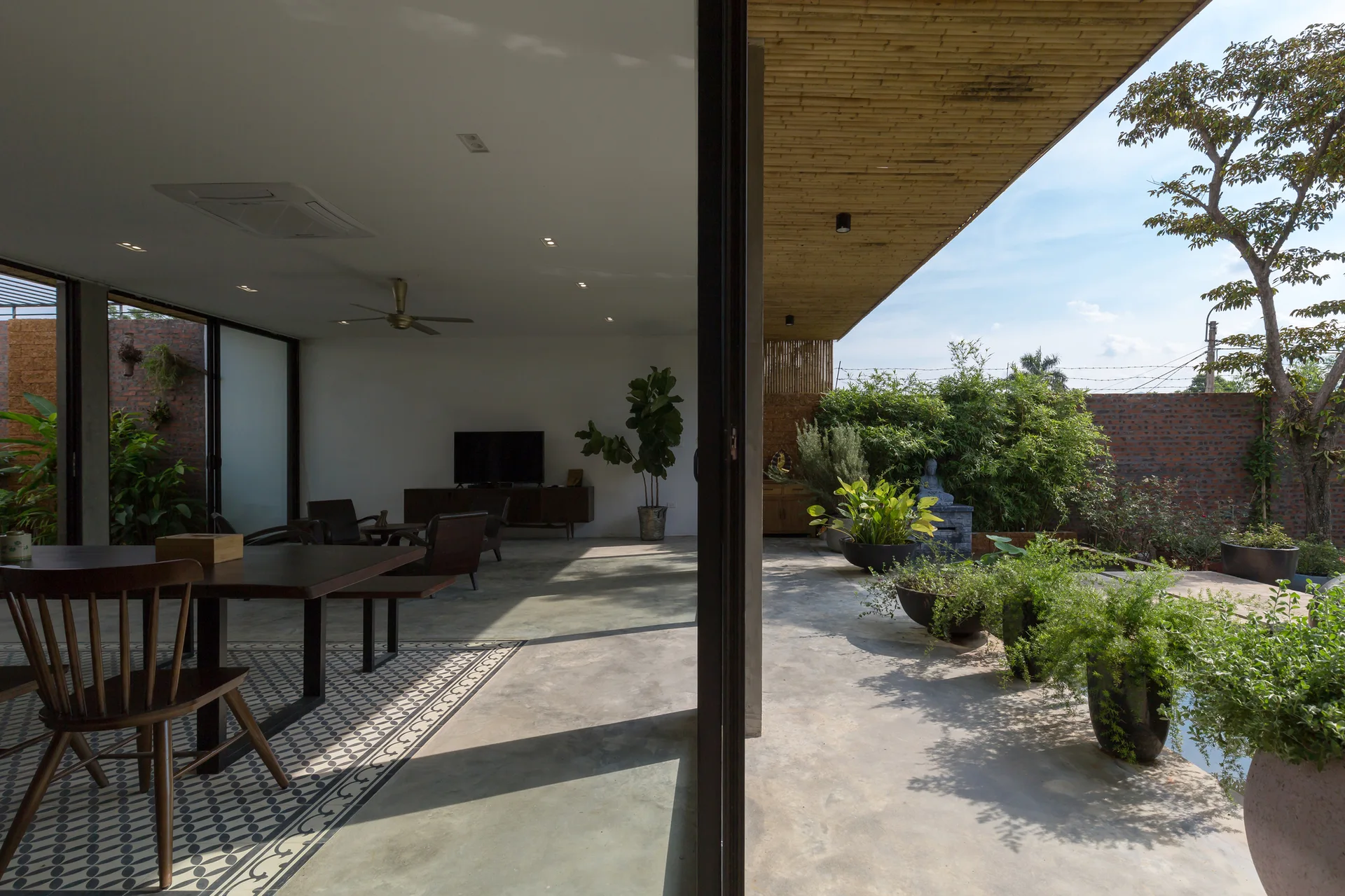 An open indoor-outdoor dining space with a wooden table on patterned floor tiles next to a polished concrete patio with lush plants.