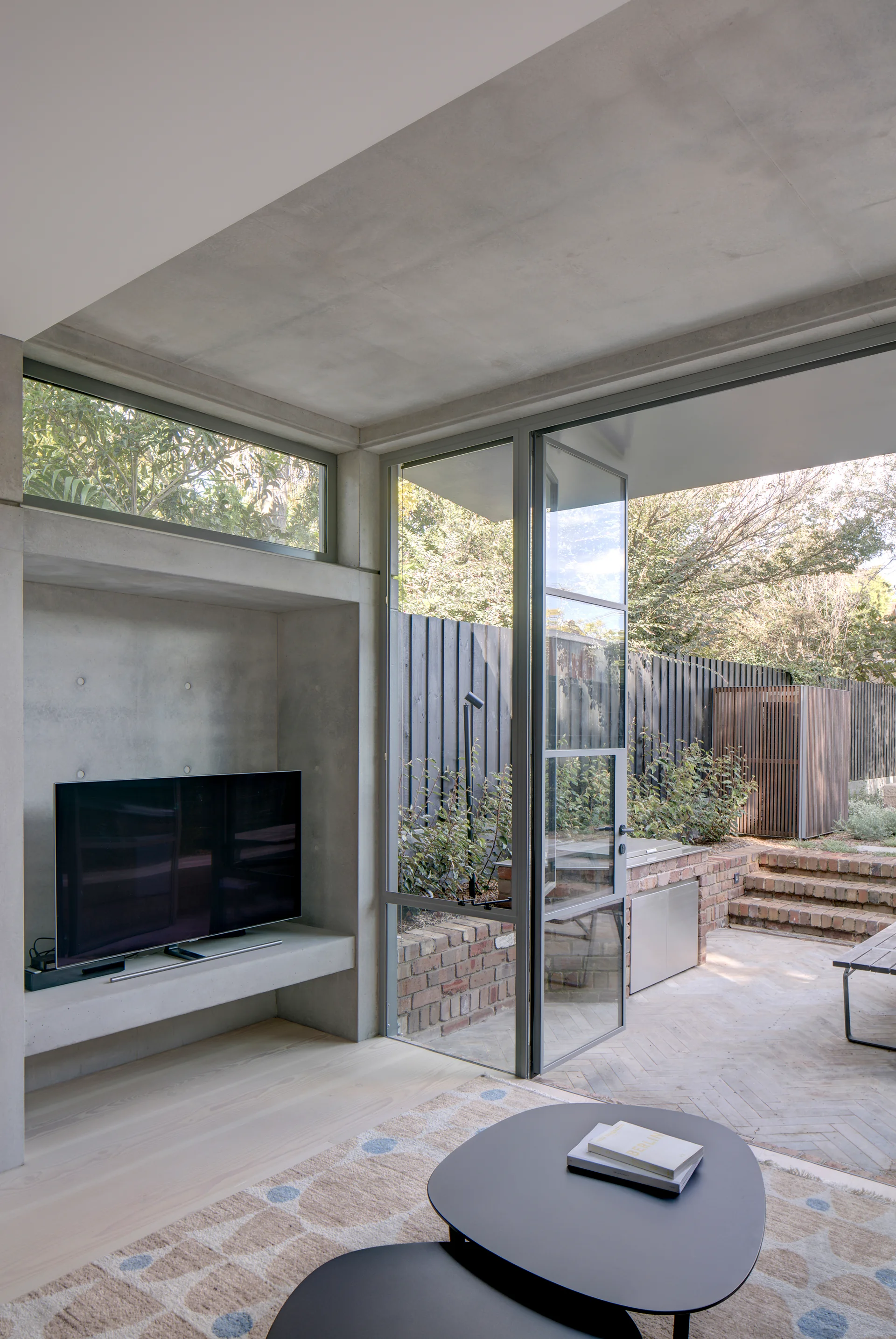 A modern living room featuring a raw concrete TV alcove, light wood floors, nesting coffee tables, and a large steel-framed glass door opening to a brick patio.