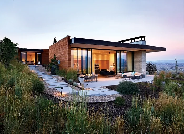 A modern hillside house featuring horizontal wood siding, a concrete patio with outdoor furniture, and a winding stone pathway through tall wild grasses.