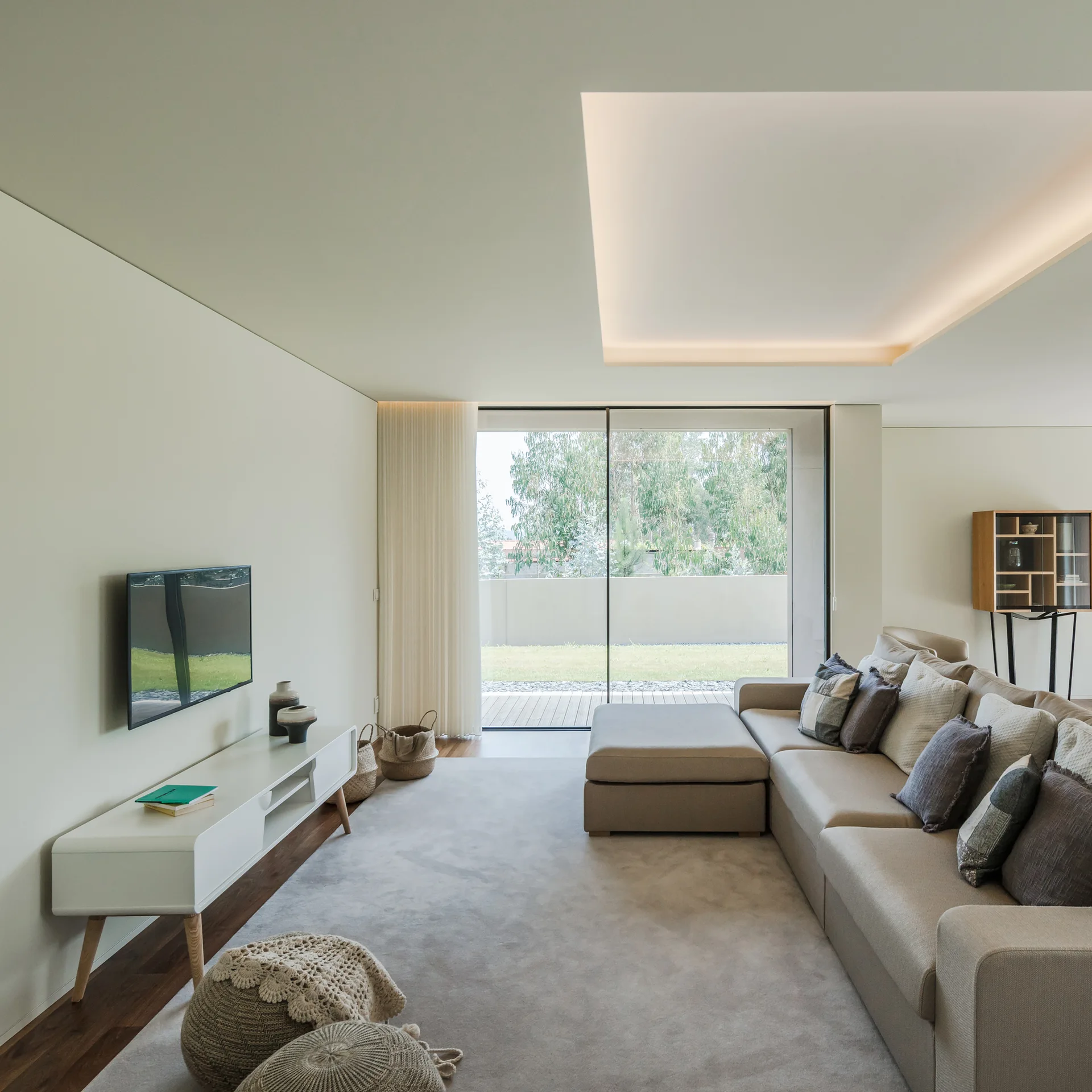A minimalist living room features a beige sectional sofa, a white media console, and sliding glass doors opening to a garden.