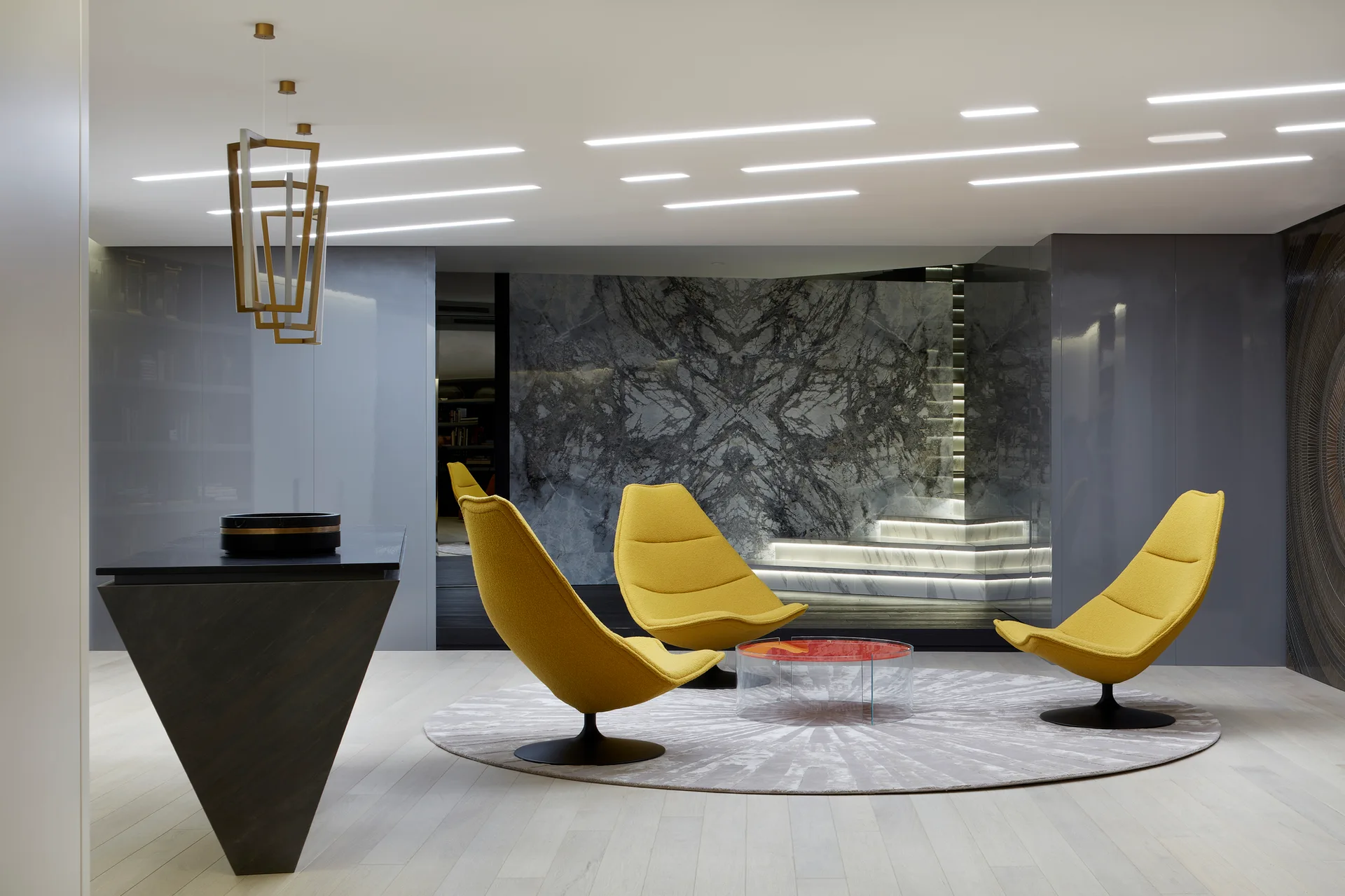 Three modern yellow lounge chairs arranged on a circular grey rug in front of a book-matched marble wall and backlit staircase.