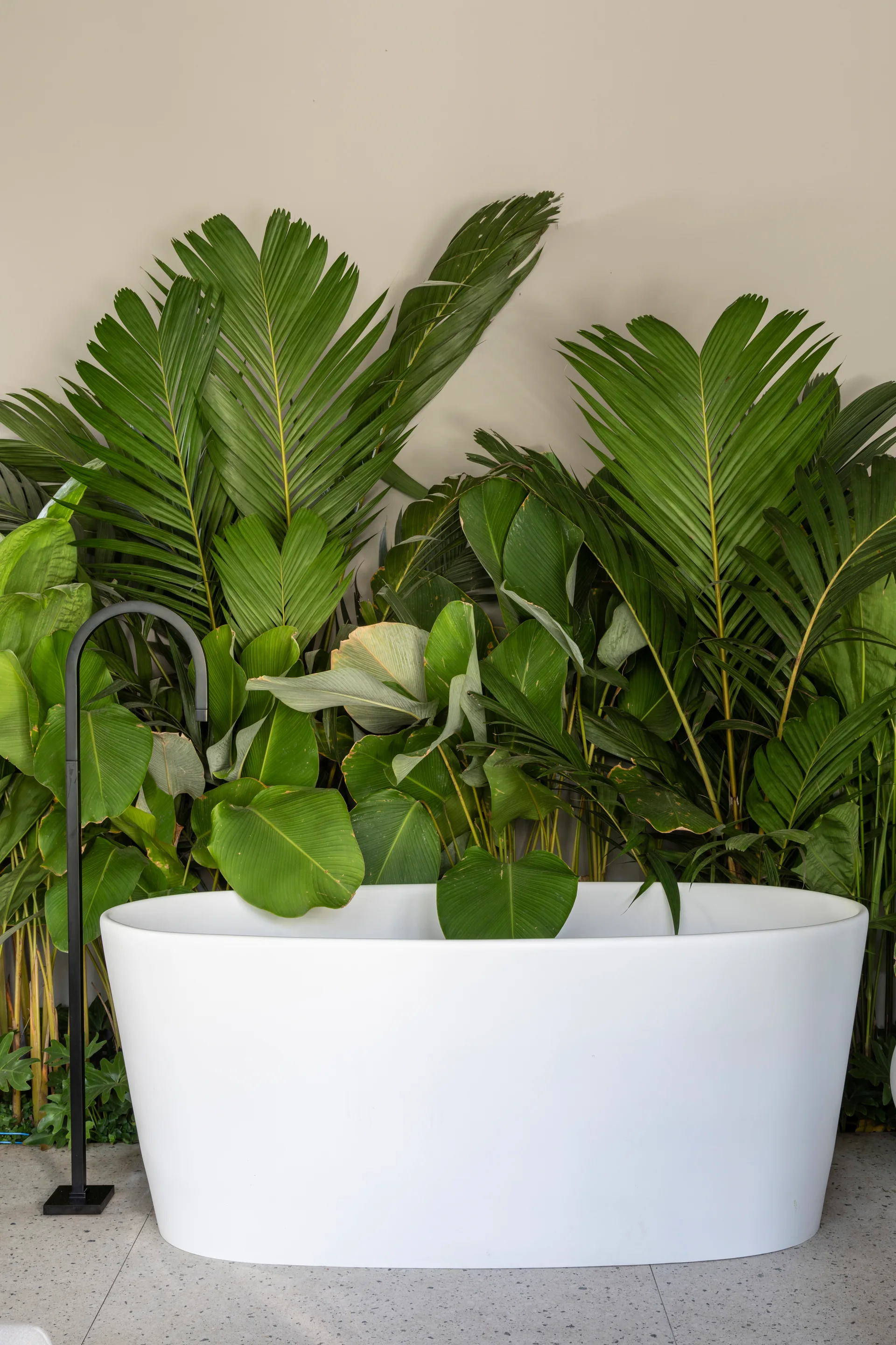 A white freestanding oval bathtub and a tall matte black faucet sit before a dense wall of lush green tropical plants on speckled grey flooring.