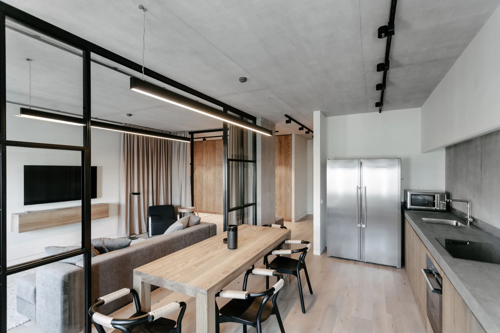 An open-concept apartment interior features a wooden dining table, black-framed glass partitions, a concrete ceiling, and a stainless steel refrigerator.