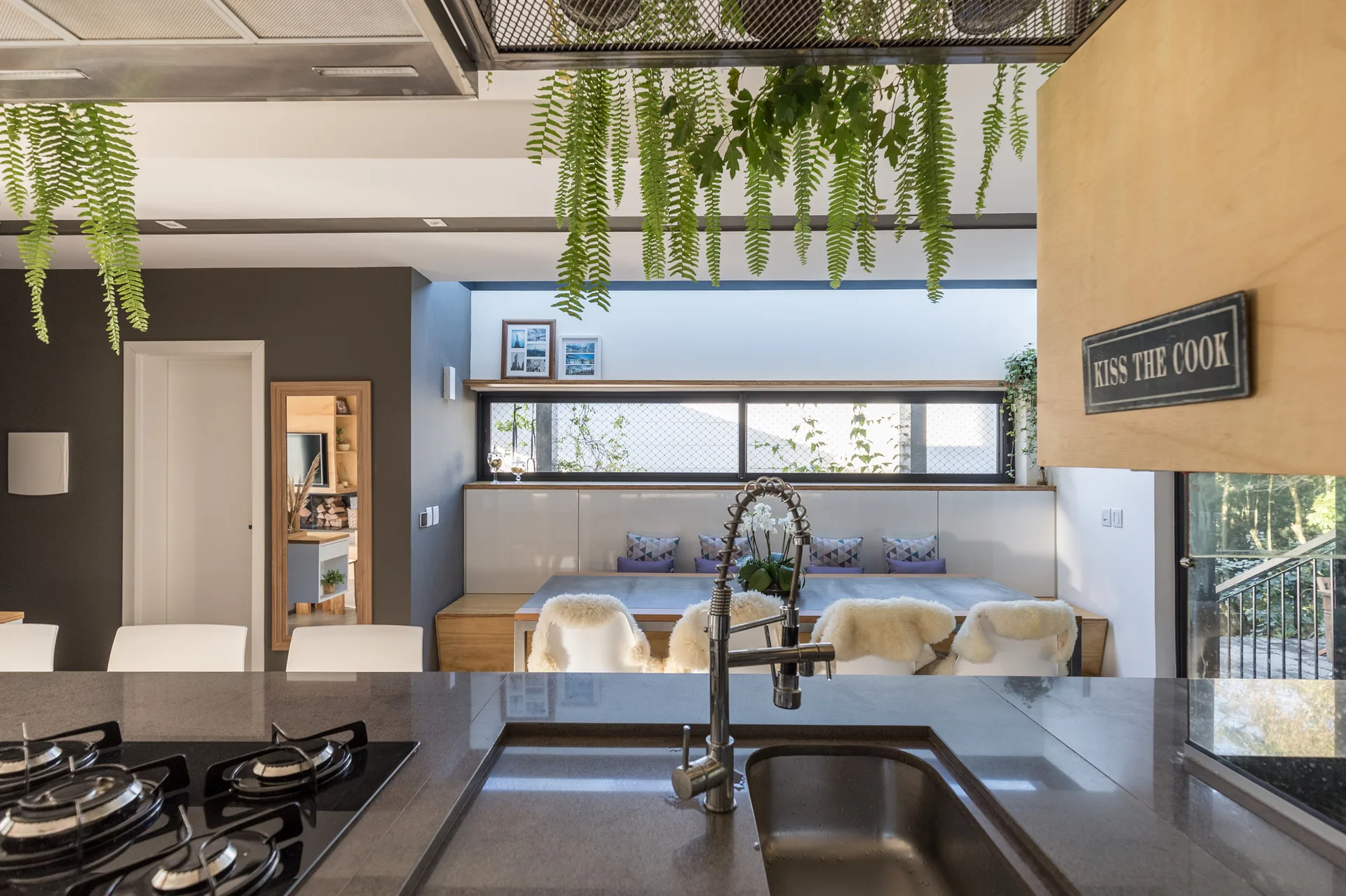A modern kitchen features a dark grey stone island with a sink and stove, looking toward a dining table with a built-in bench under a ribbon window.