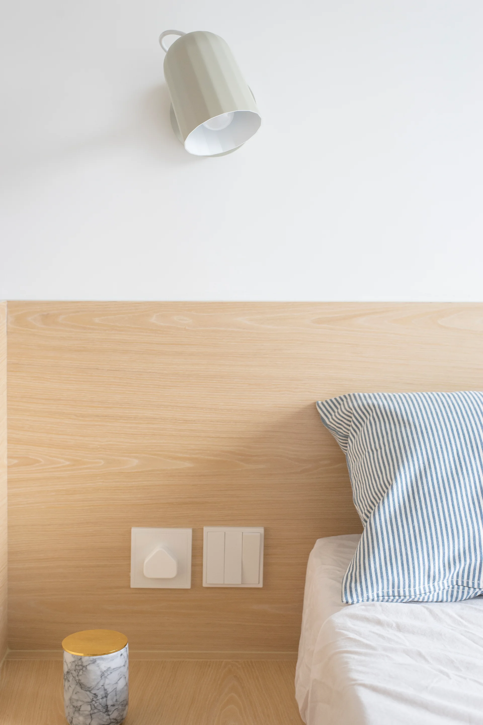 A close-up of a light wood headboard panel with white switches, a blue striped pillow, and a sage green wall sconce mounted on a white wall.
