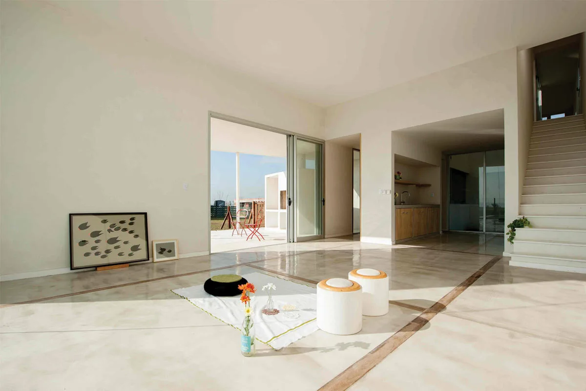 A minimalist open-plan living room features polished concrete floors with wood inlays, a white staircase, and sliding glass doors opening to a patio.