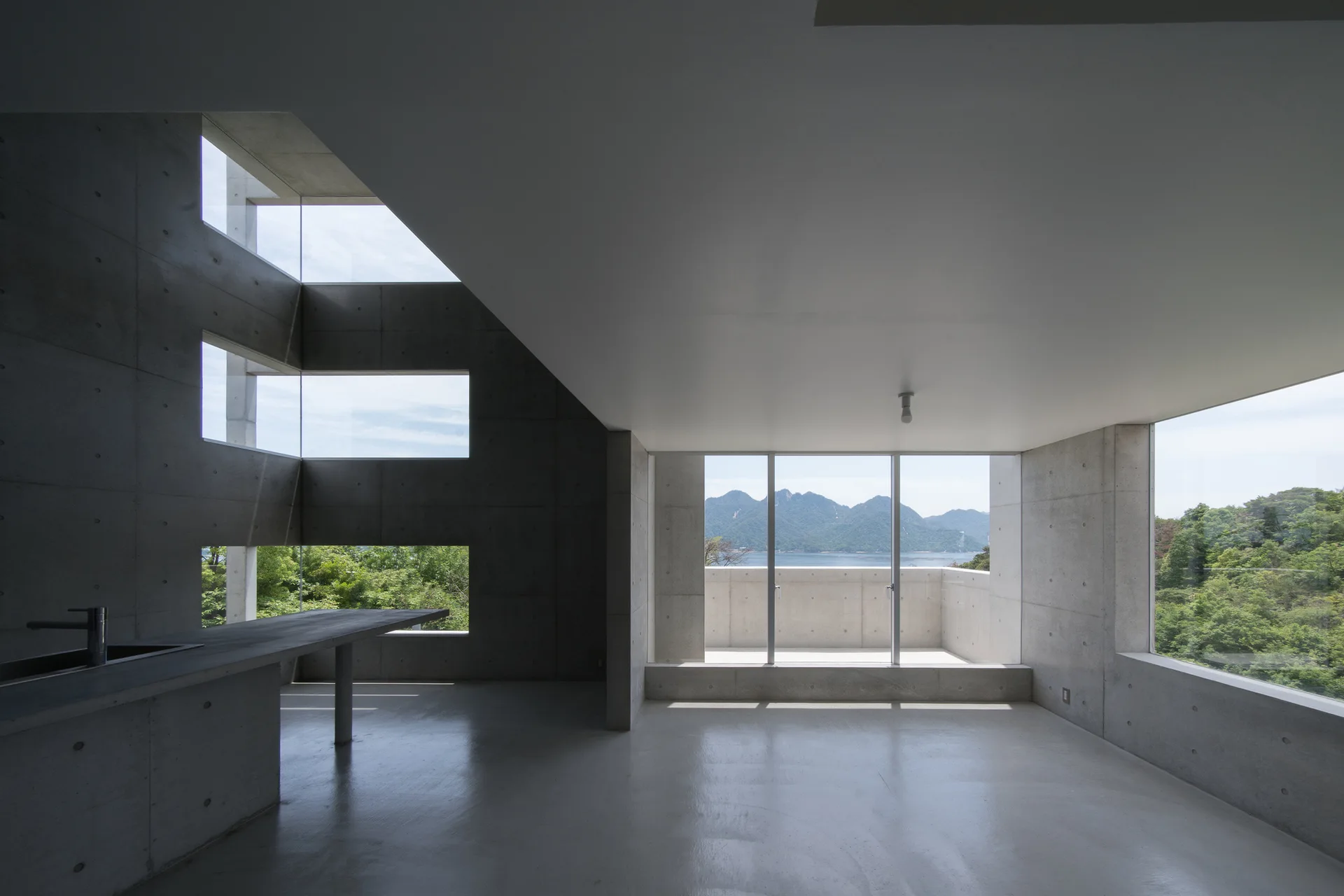 A minimalist concrete interior features a grey kitchen island, smooth concrete walls with tie-rod holes, and large glass windows overlooking mountains.