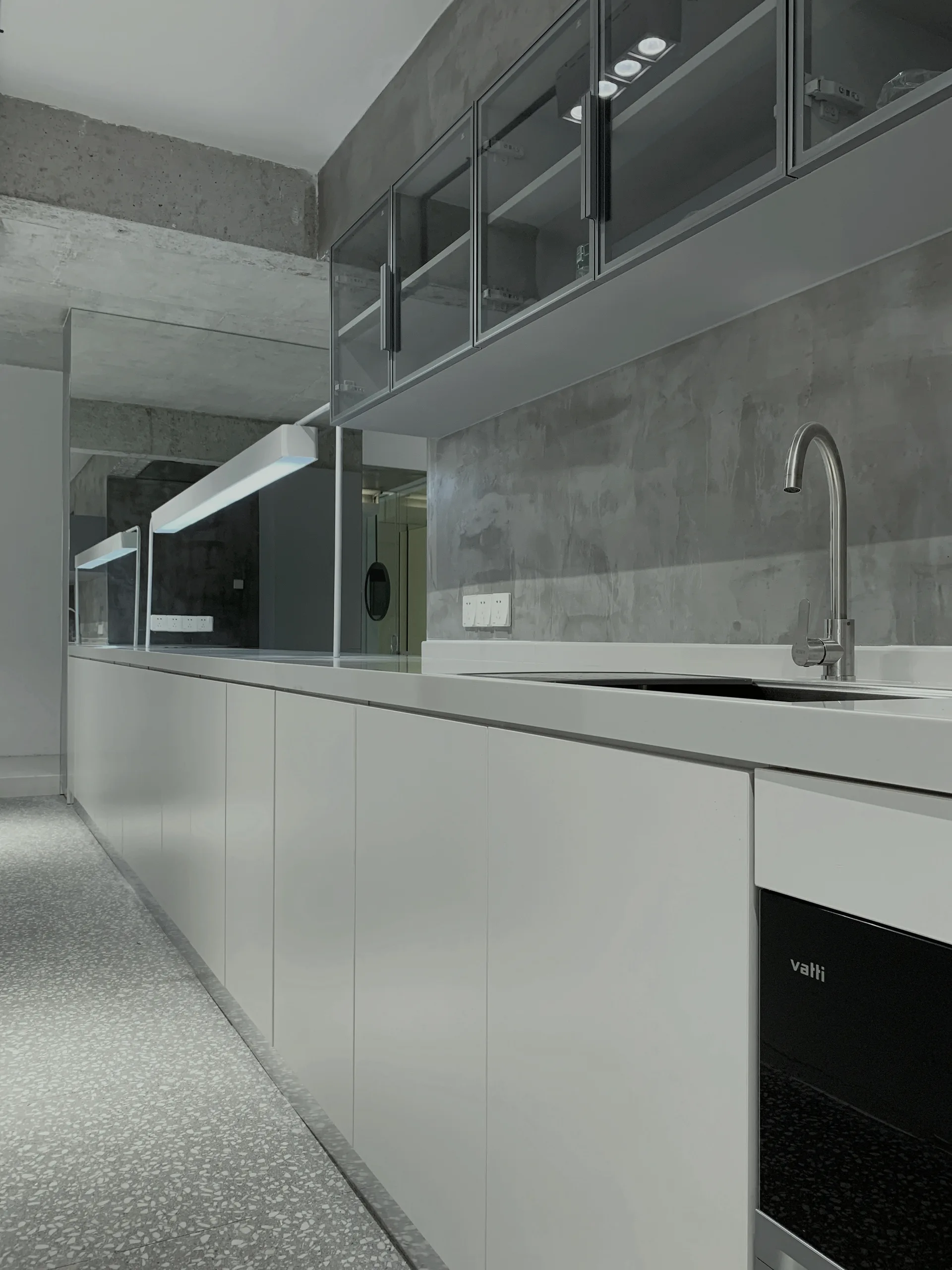 A minimalist kitchen features seamless white lower cabinetry, glass-fronted upper cupboards, raw concrete walls, and speckled terrazzo flooring.