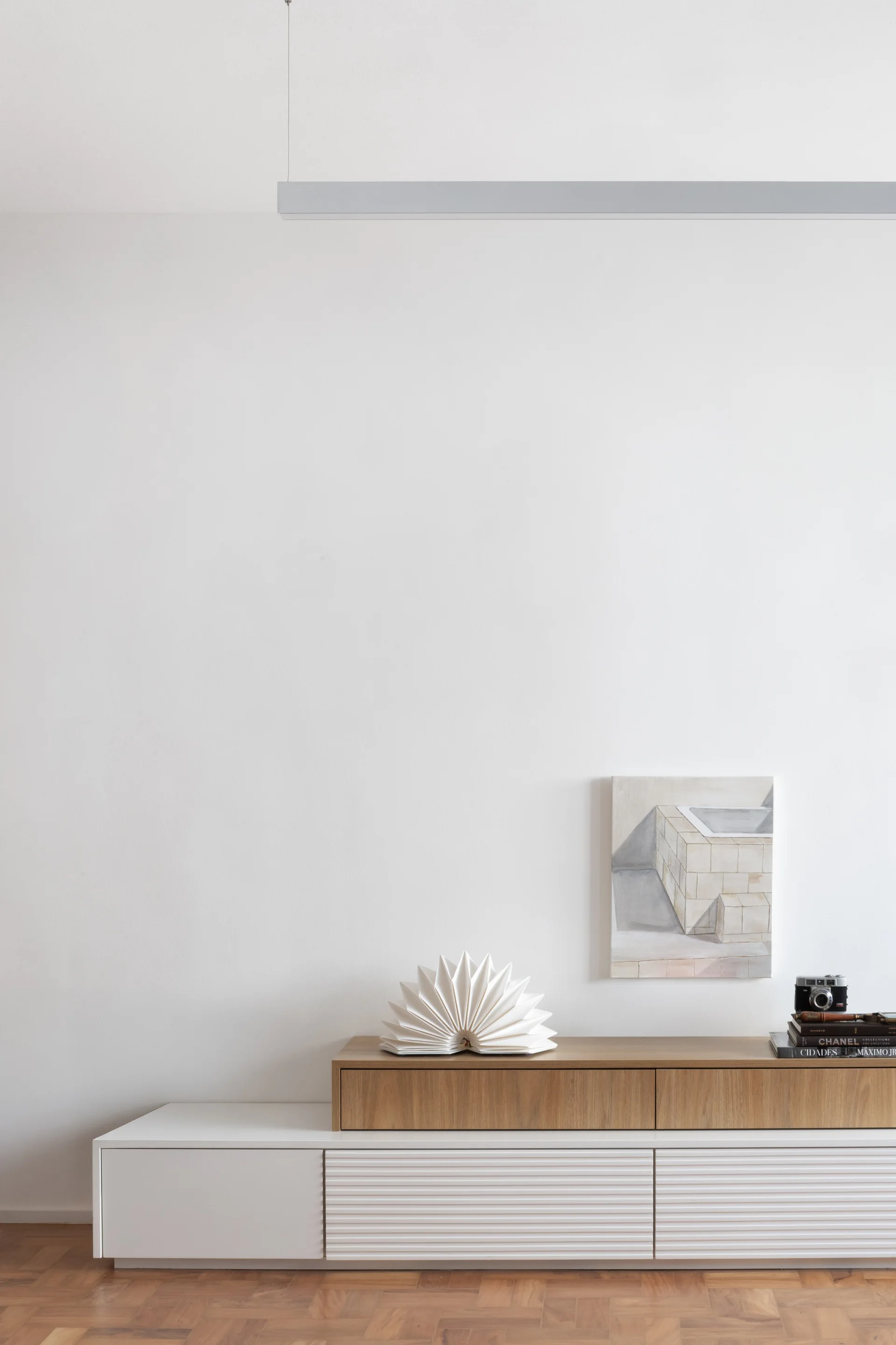 A minimalist wood and white media console with slatted doors sits against a white wall with a small painting and paper sculpture.
