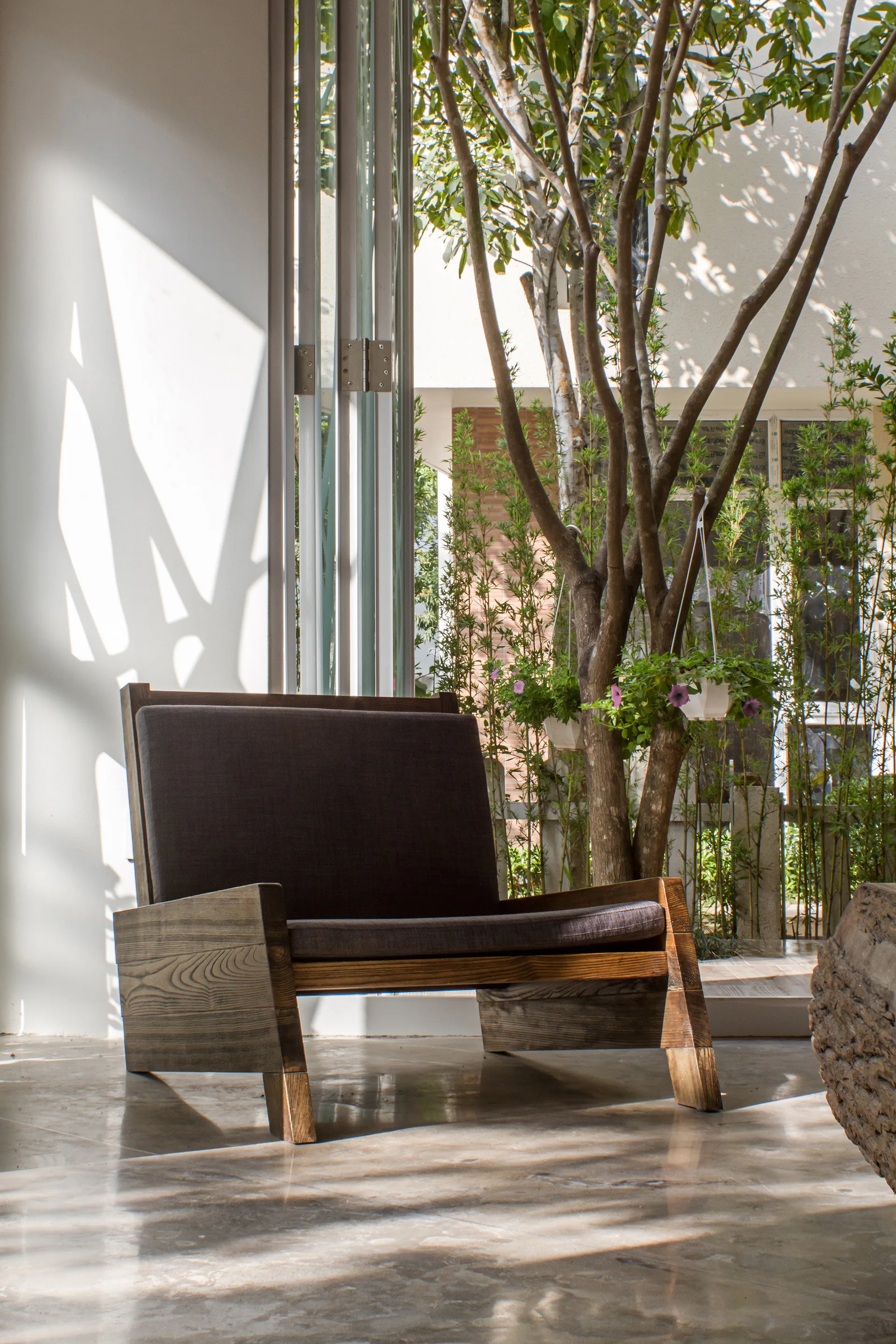 A low-profile wooden armchair with a dark cushion sits on a polished floor near open bi-fold glass doors leading to a green garden.