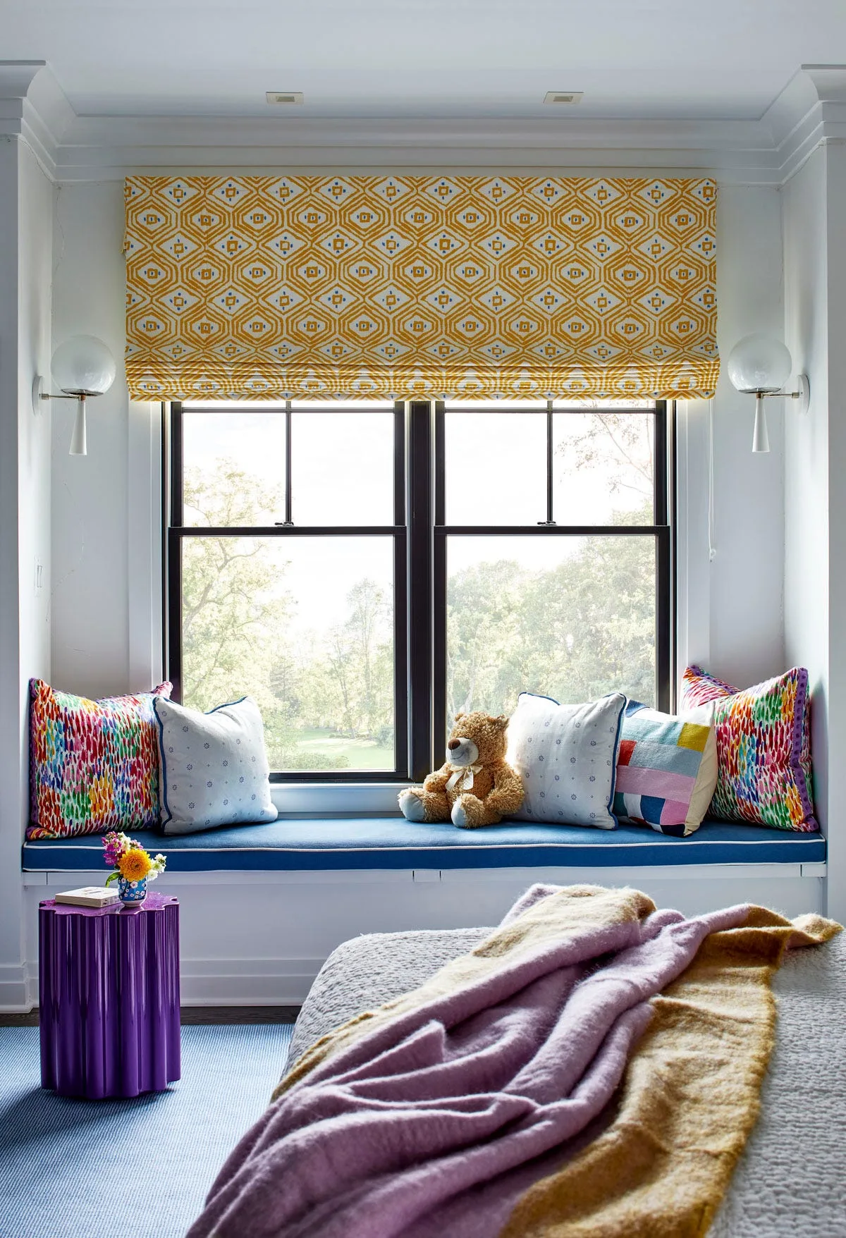 A bedroom window seat features a blue cushion, colorful pillows, and a patterned yellow roman shade next to a fluted purple side table.