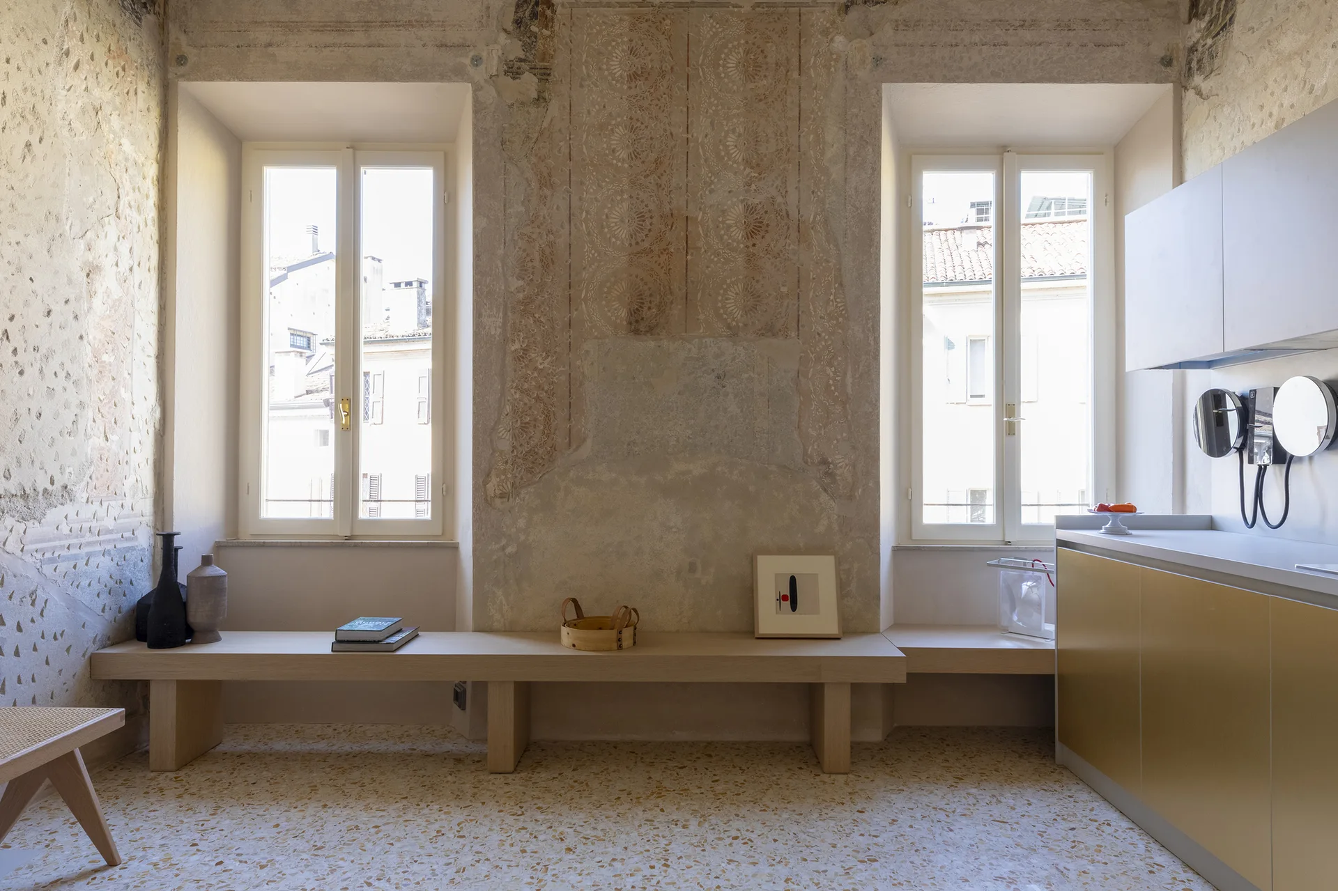 A long light wood bench sits below two tall windows in a kitchen featuring distressed plaster walls, terrazzo flooring, and metallic gold cabinets.