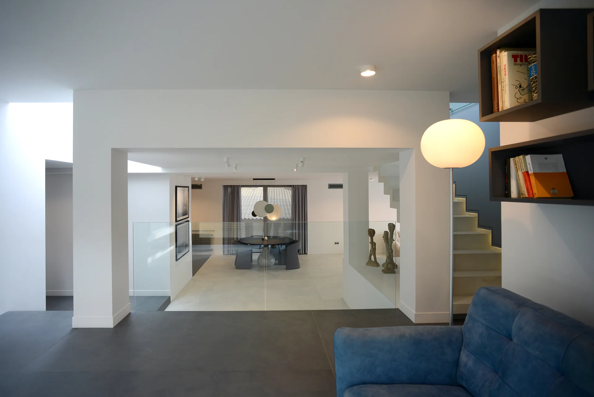 A modern split-level interior featuring a blue sofa and globe floor lamp in the foreground with a dining area visible below behind a glass railing.