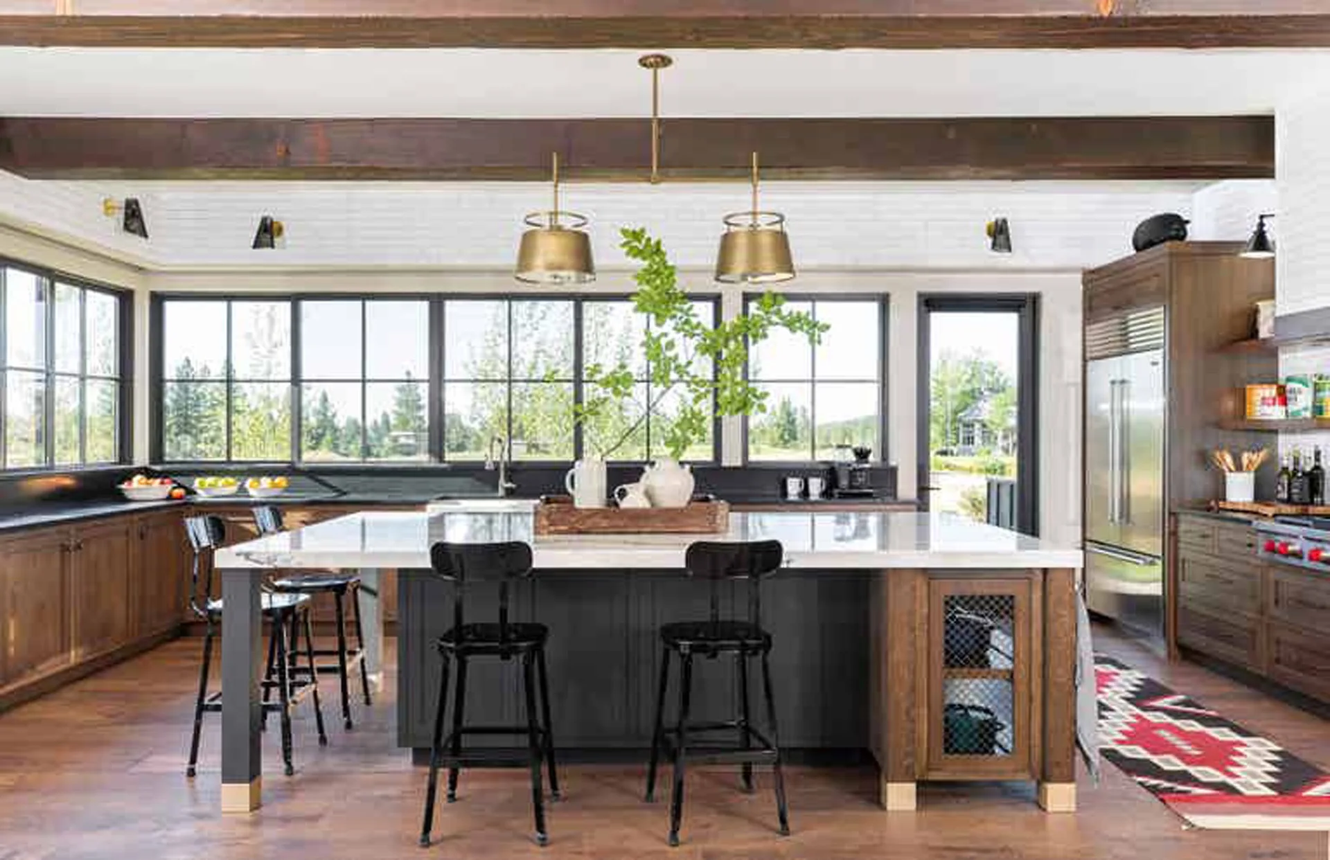 A spacious kitchen with a white-topped wooden island, black bar stools, dark wood ceiling beams, and large black-framed windows overlooking trees.