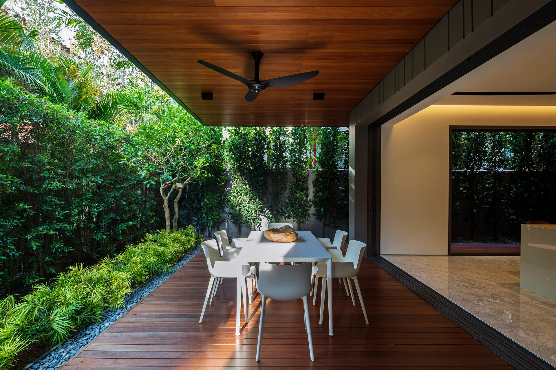 A modern covered patio with a white dining set on a wood plank deck, bordered by lush green landscaping and a large glass sliding door.