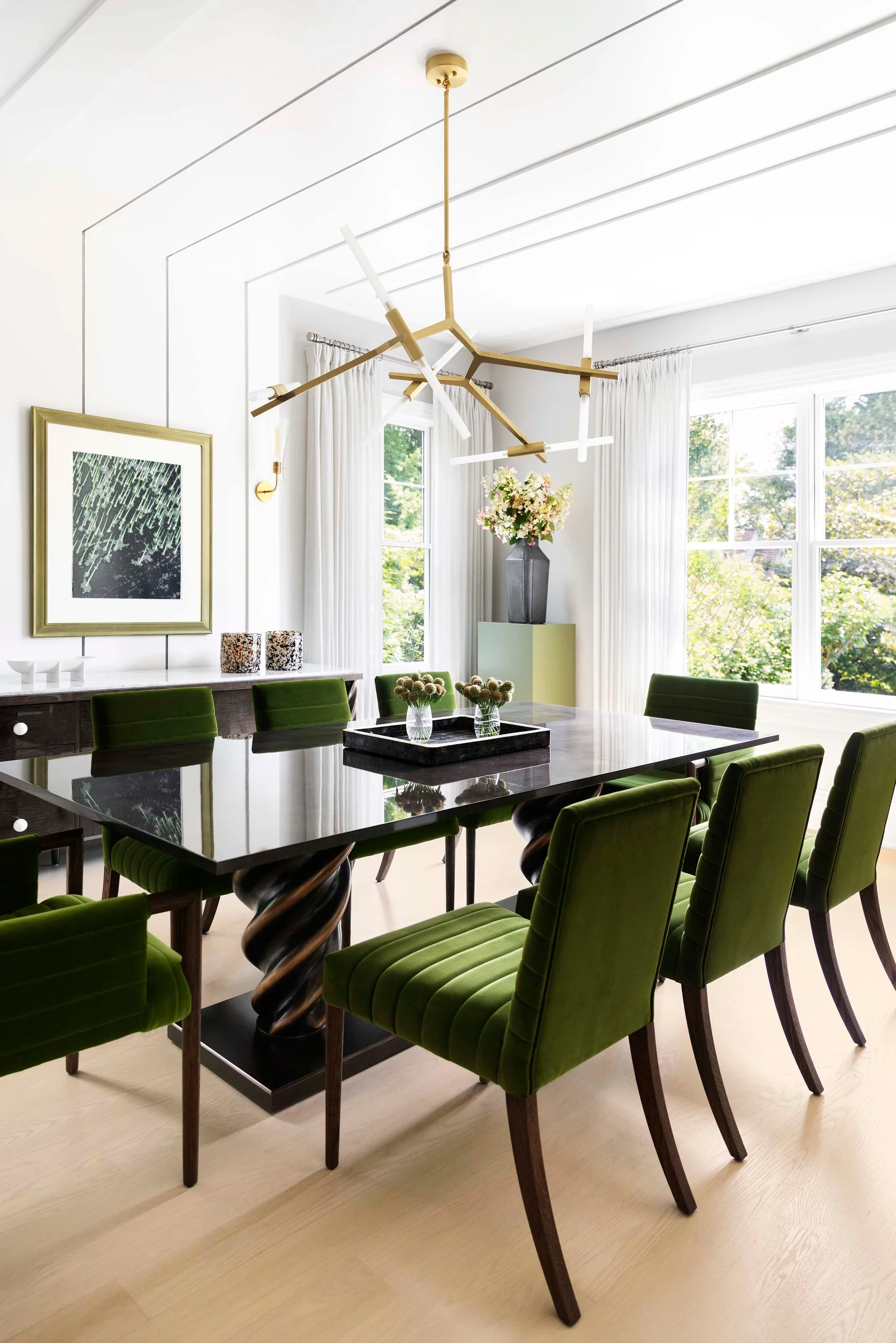 Green velvet chairs surround a dark glossy dining table with a twisted spiral base beneath a geometric brass chandelier.