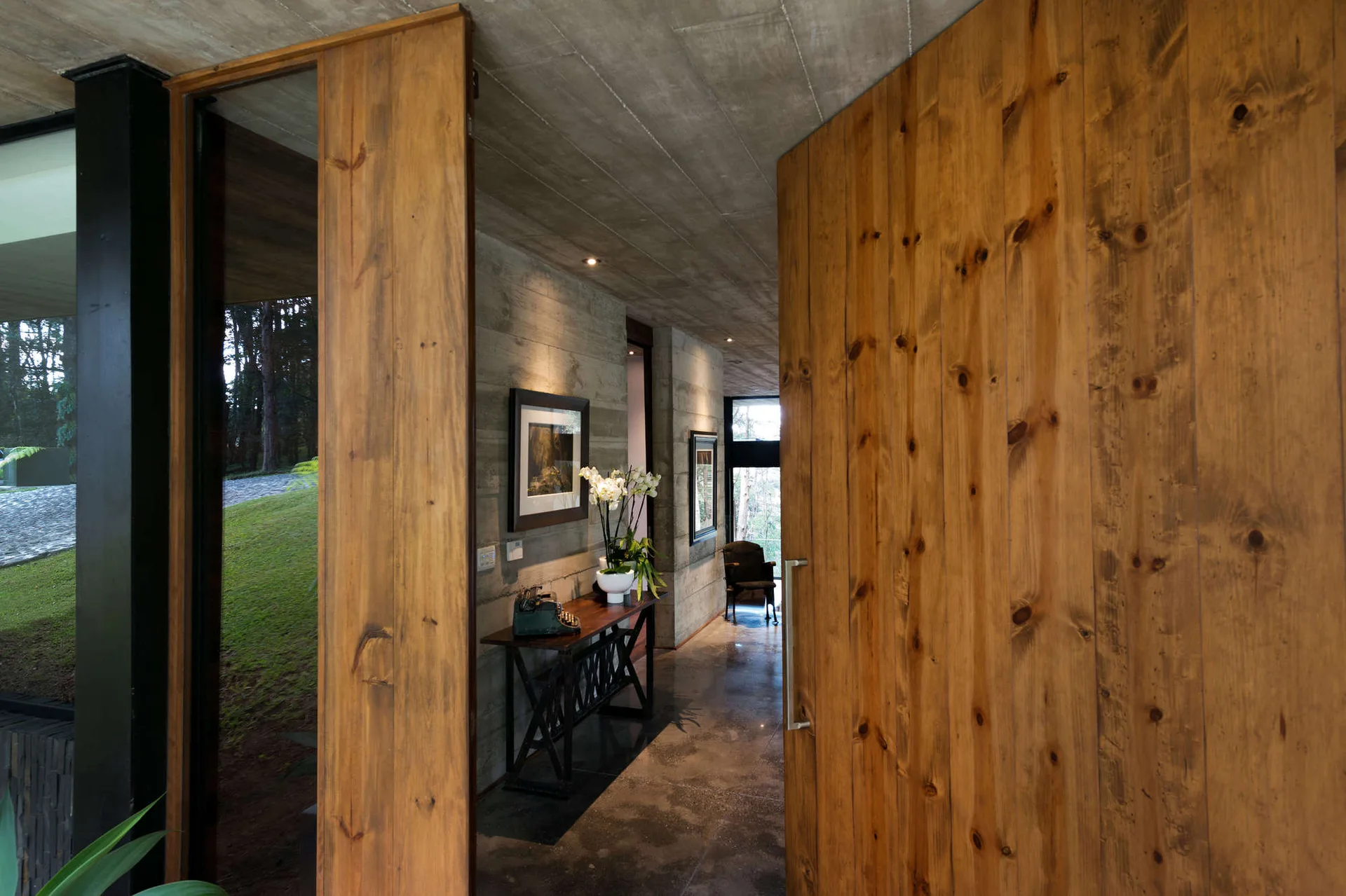 An open wooden pivot door leads into a concrete entryway hallway with board-formed walls and a dark wood console table.