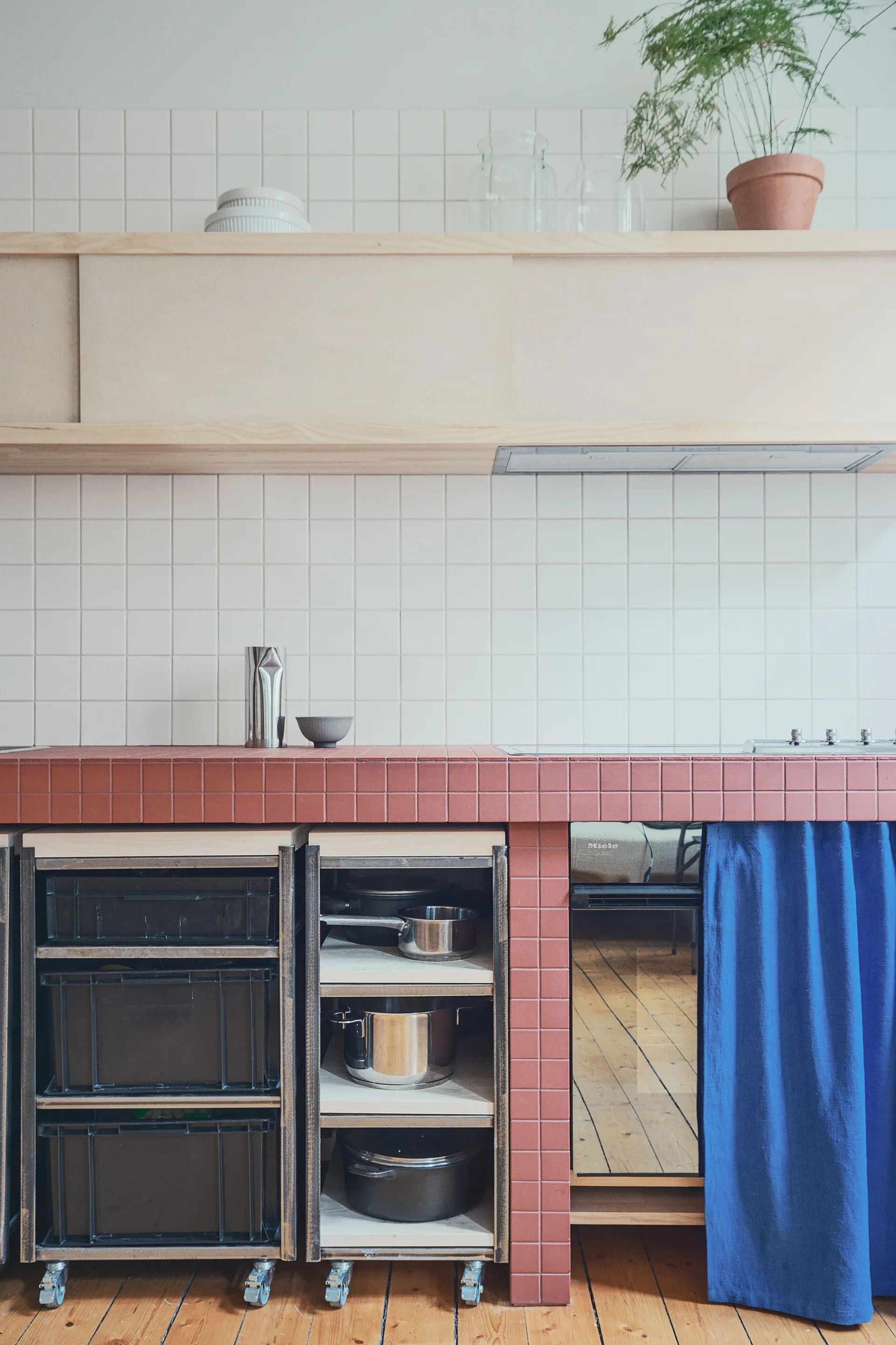 A modern kitchen with a red tiled countertop, white grid backsplash, mobile wooden storage carts on casters, and a bright blue fabric curtain.