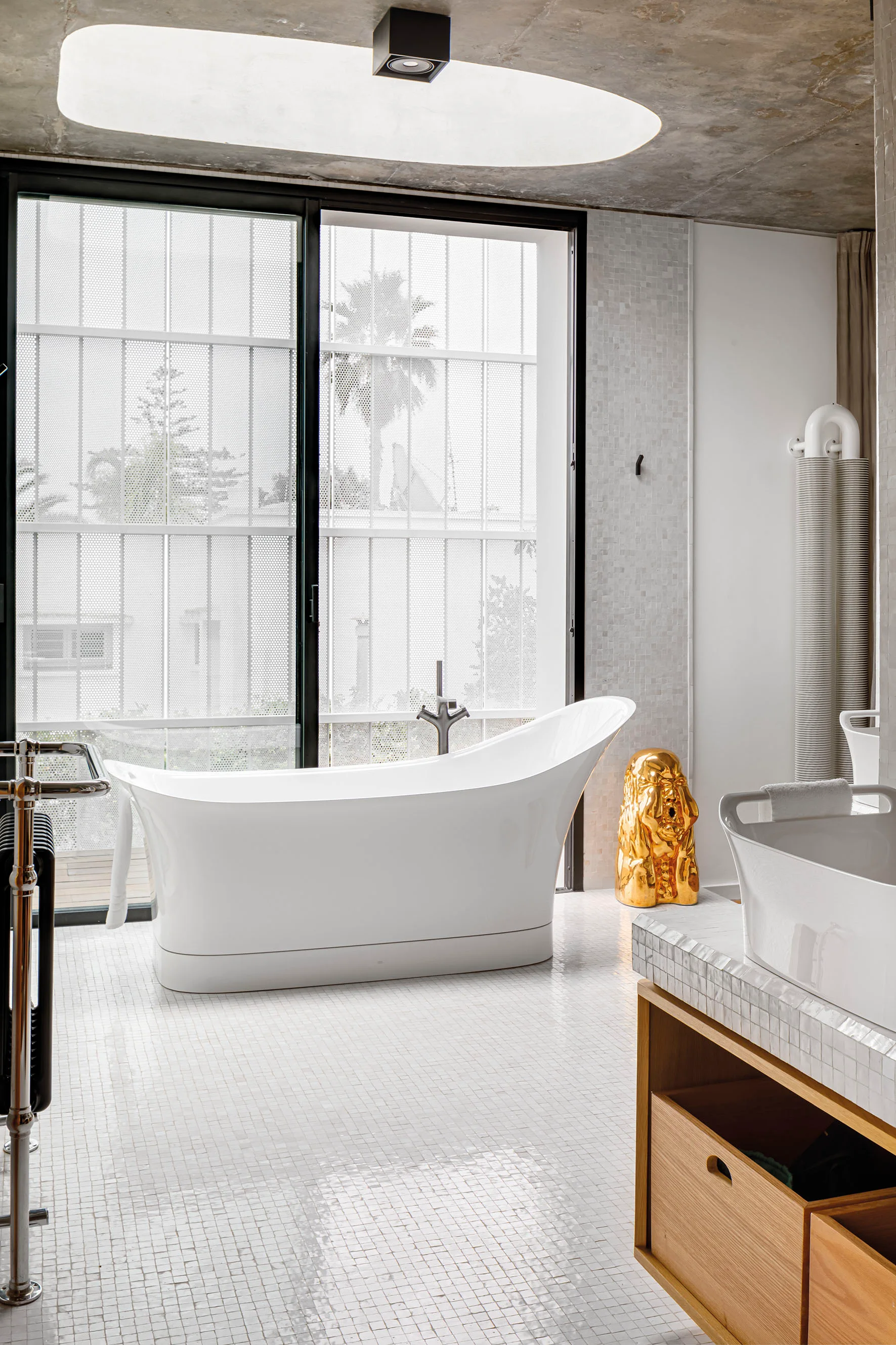 A modern bathroom features a white freestanding bathtub on small mosaic tiles under an exposed concrete ceiling with a skylight.