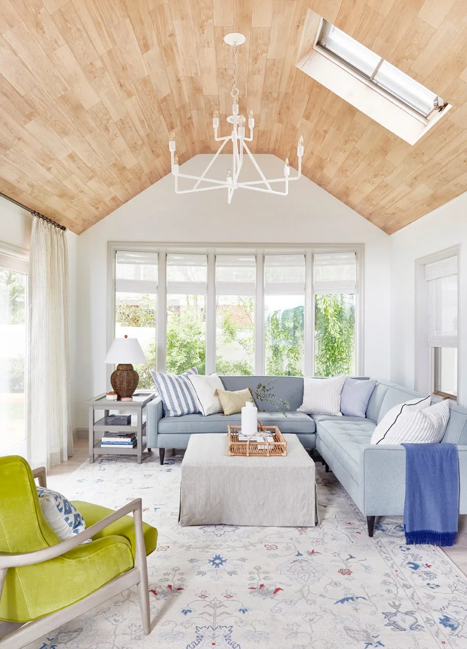 A light-filled living room featuring a blue sectional, a lime green armchair, and a vaulted wood-paneled ceiling with a skylight and a white chandelier.