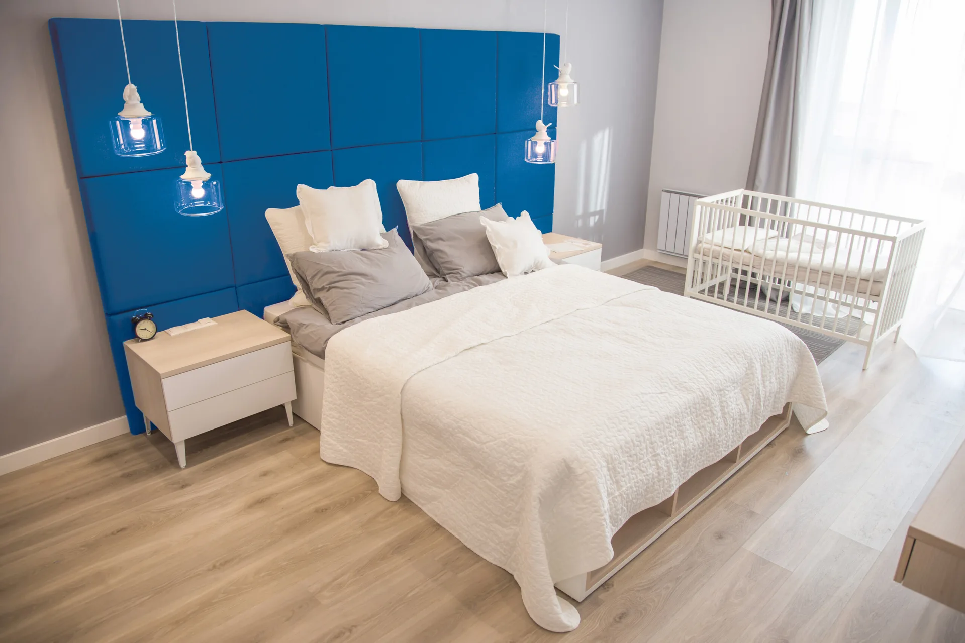 A gray bedroom features a bed with a large blue upholstered grid headboard, matching white nightstands, pendant lights, and a white wooden baby crib.
