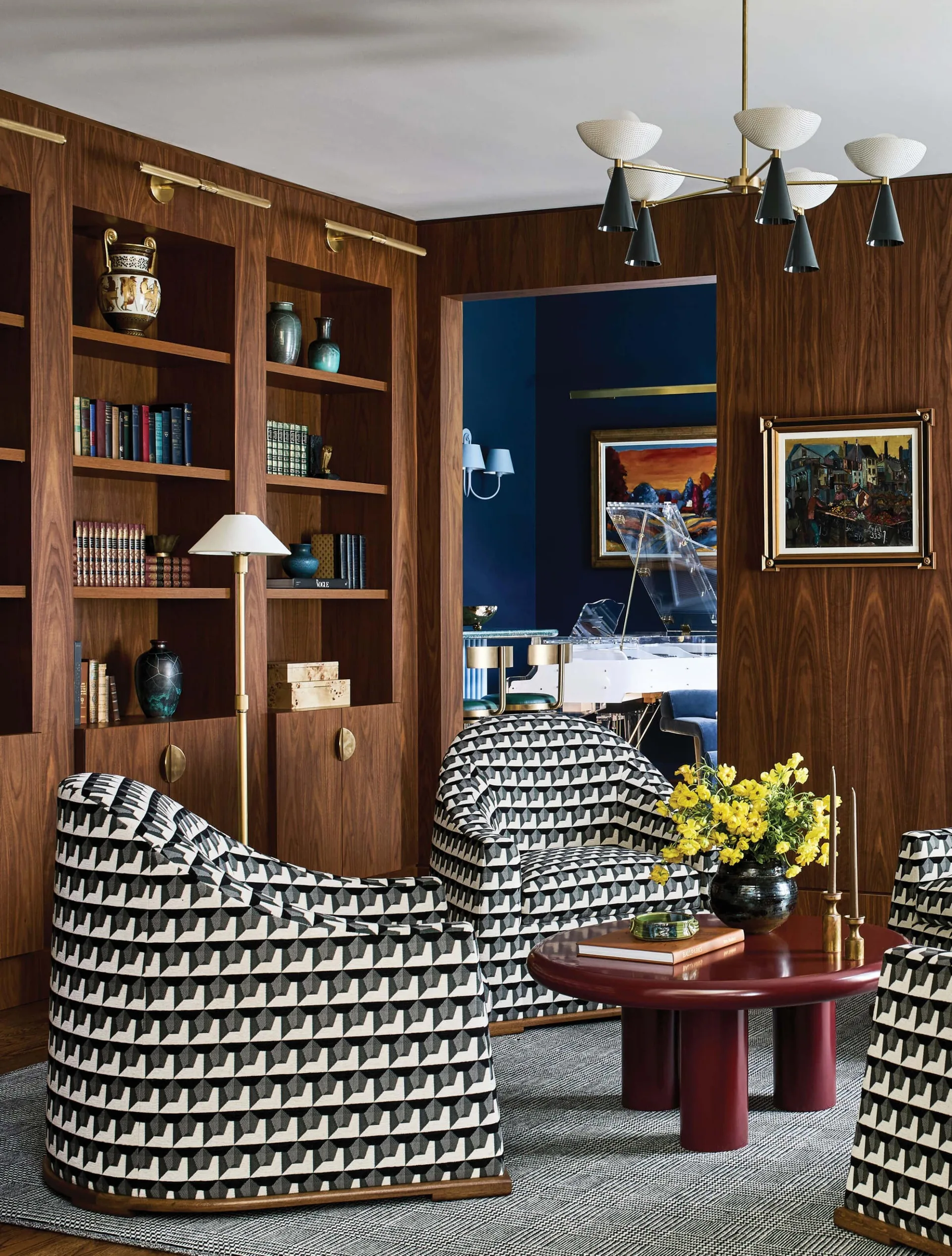 A wood-paneled living room features black and white geometric patterned armchairs, a round burgundy coffee table, and bookshelves.