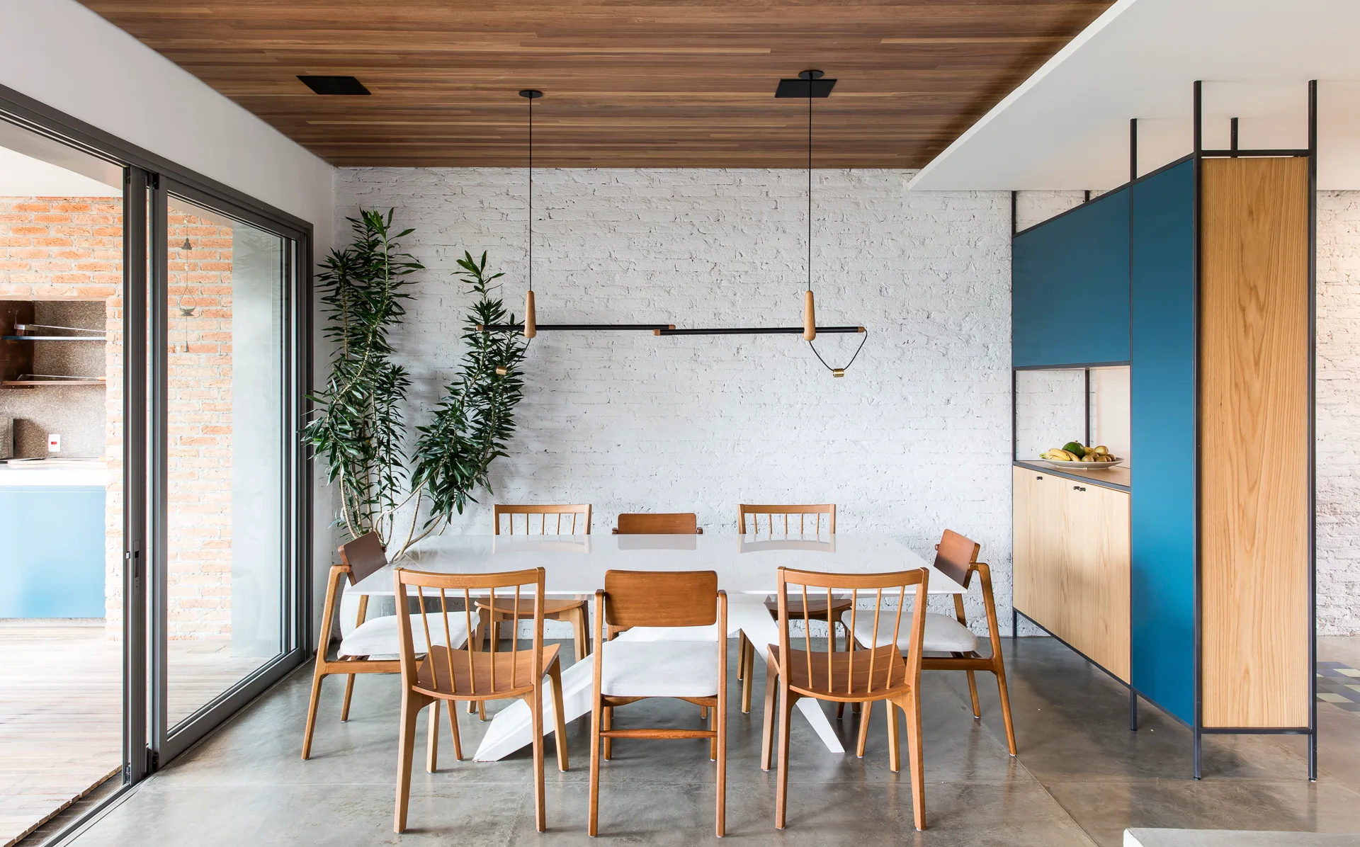 A modern dining room features a white table and wood chairs set against a white painted brick wall under a wood plank ceiling.
