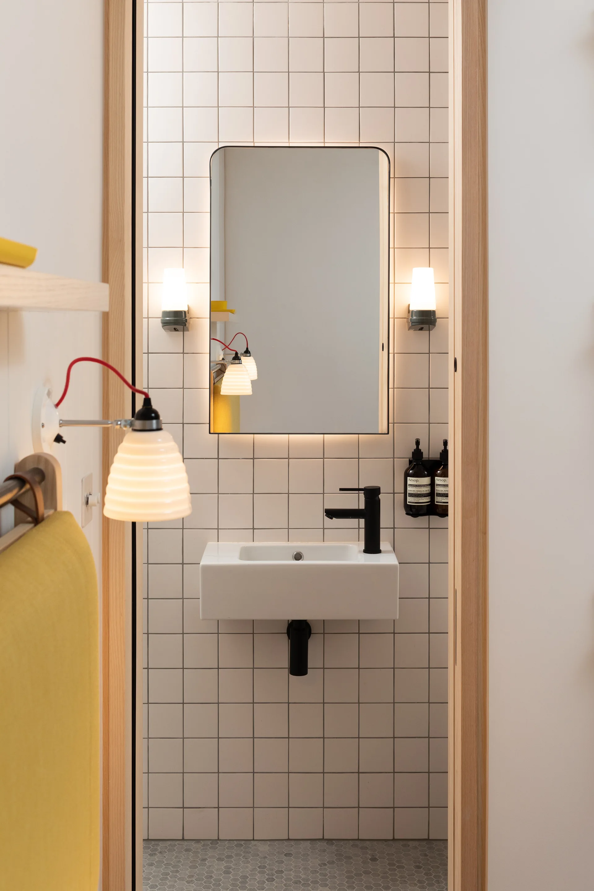 A narrow powder room viewed through a wooden doorway, featuring white grid-tiled walls, a wall-mounted sink with a black faucet, and a backlit mirror.