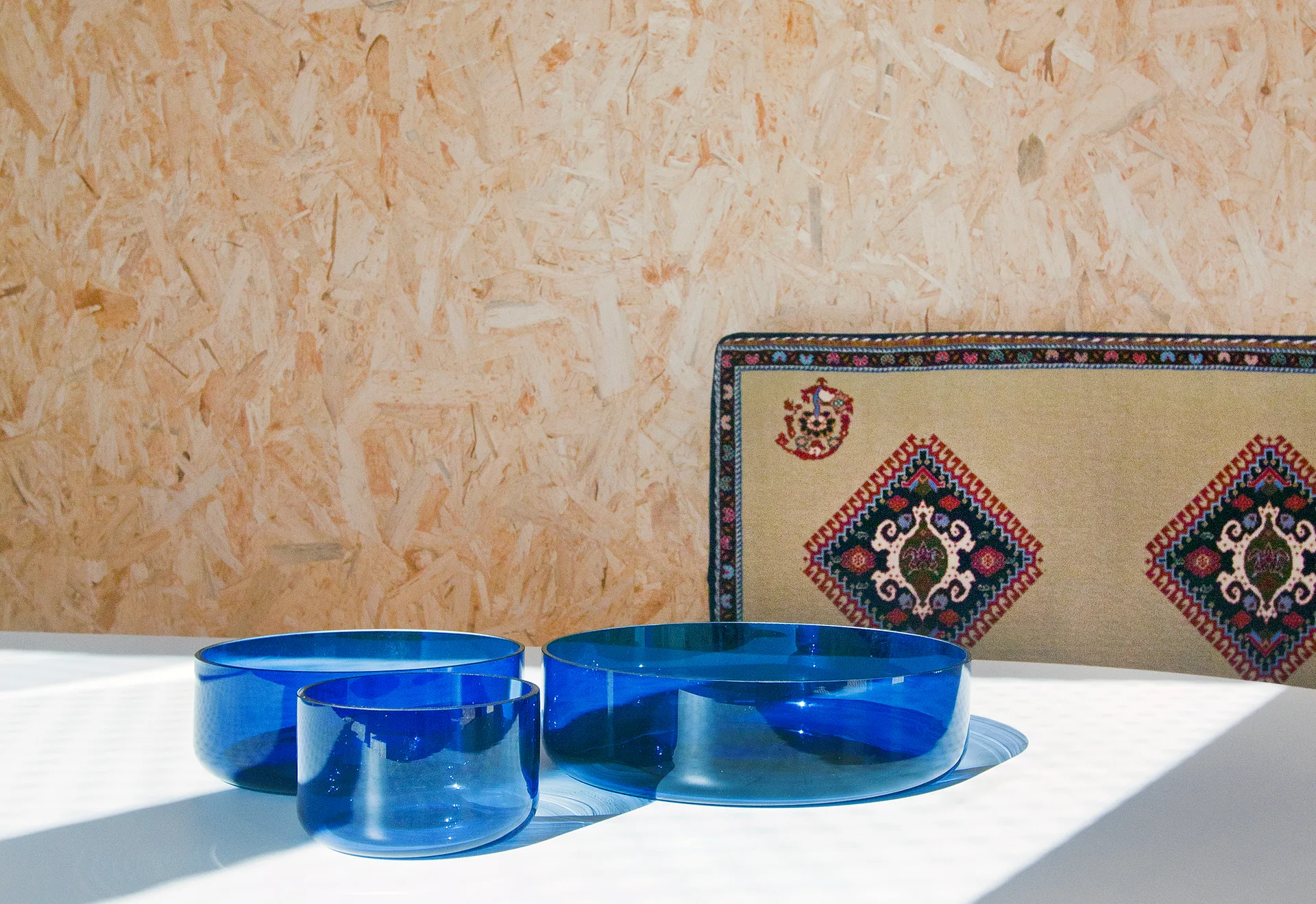 Three translucent blue glass bowls sit on a white table in front of an oriented strand board wall and a patterned geometric textile.