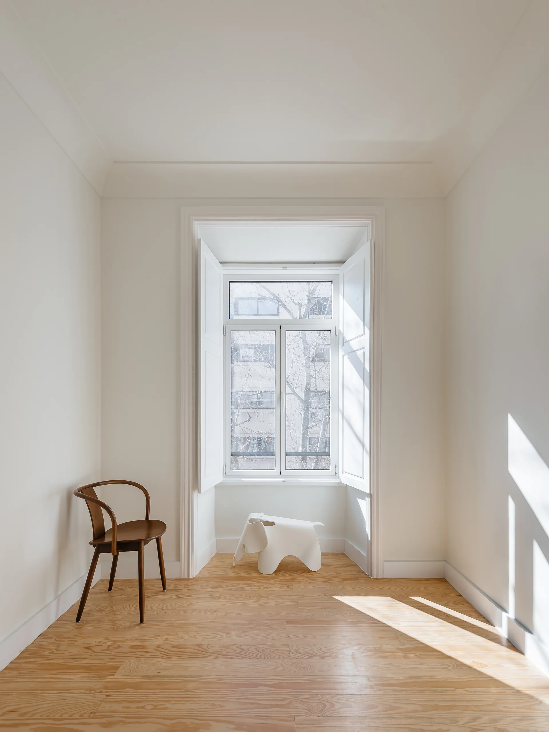 A minimalist room corner features a recessed window with white shutters, light wood floors, a wooden chair, and a white elephant stool.