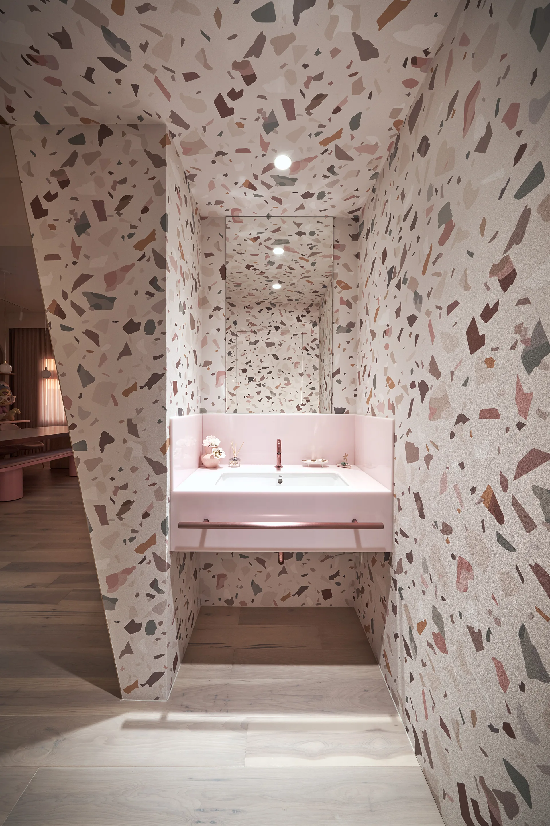 A modern powder room featuring pink terrazzo walls and ceiling, a custom pink wall-mounted sink with copper hardware, and a tall mirror.