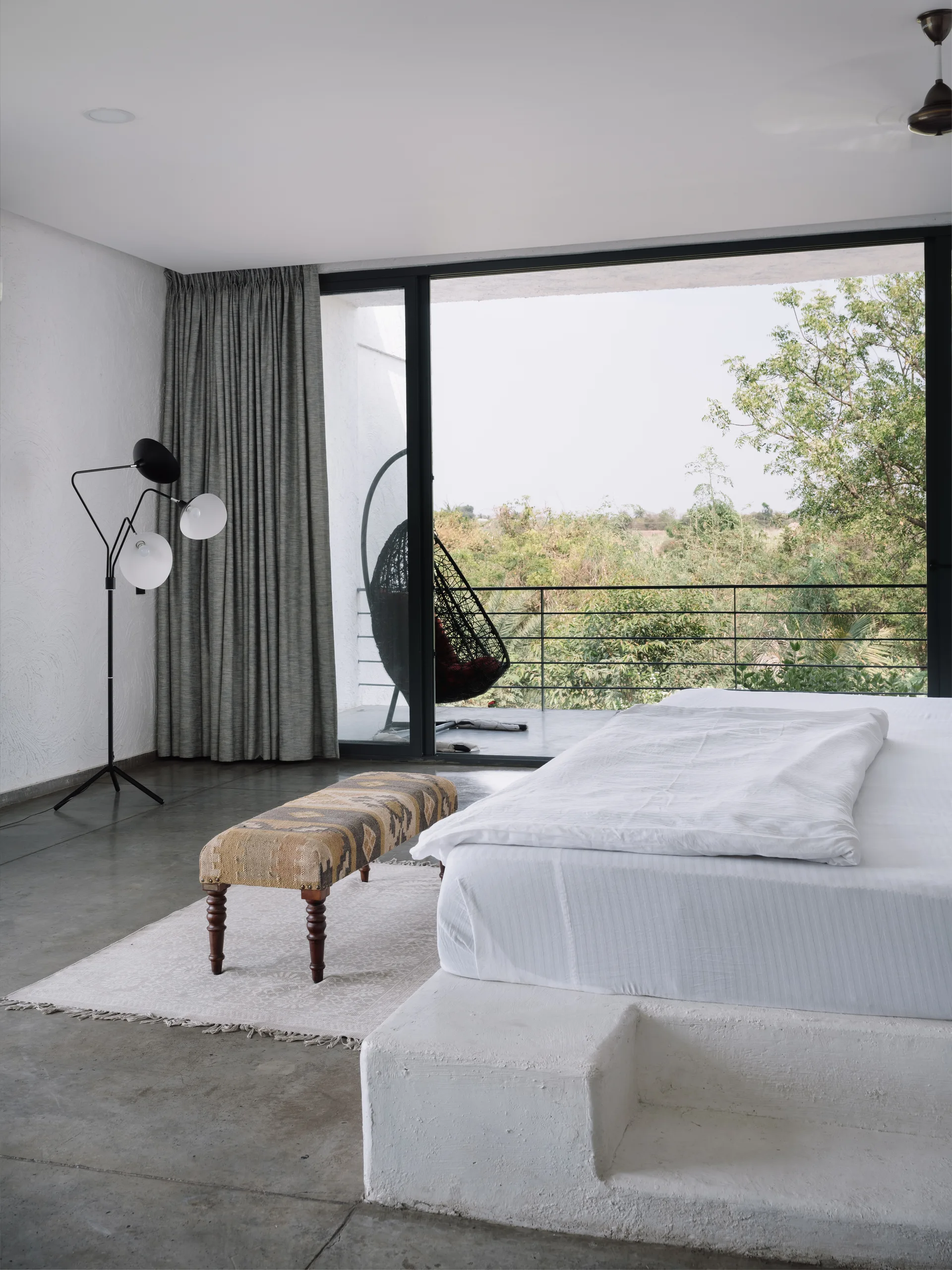 A minimalist bedroom features a built-in white plaster platform bed, gray concrete floors, and sliding glass doors opening to a balcony.