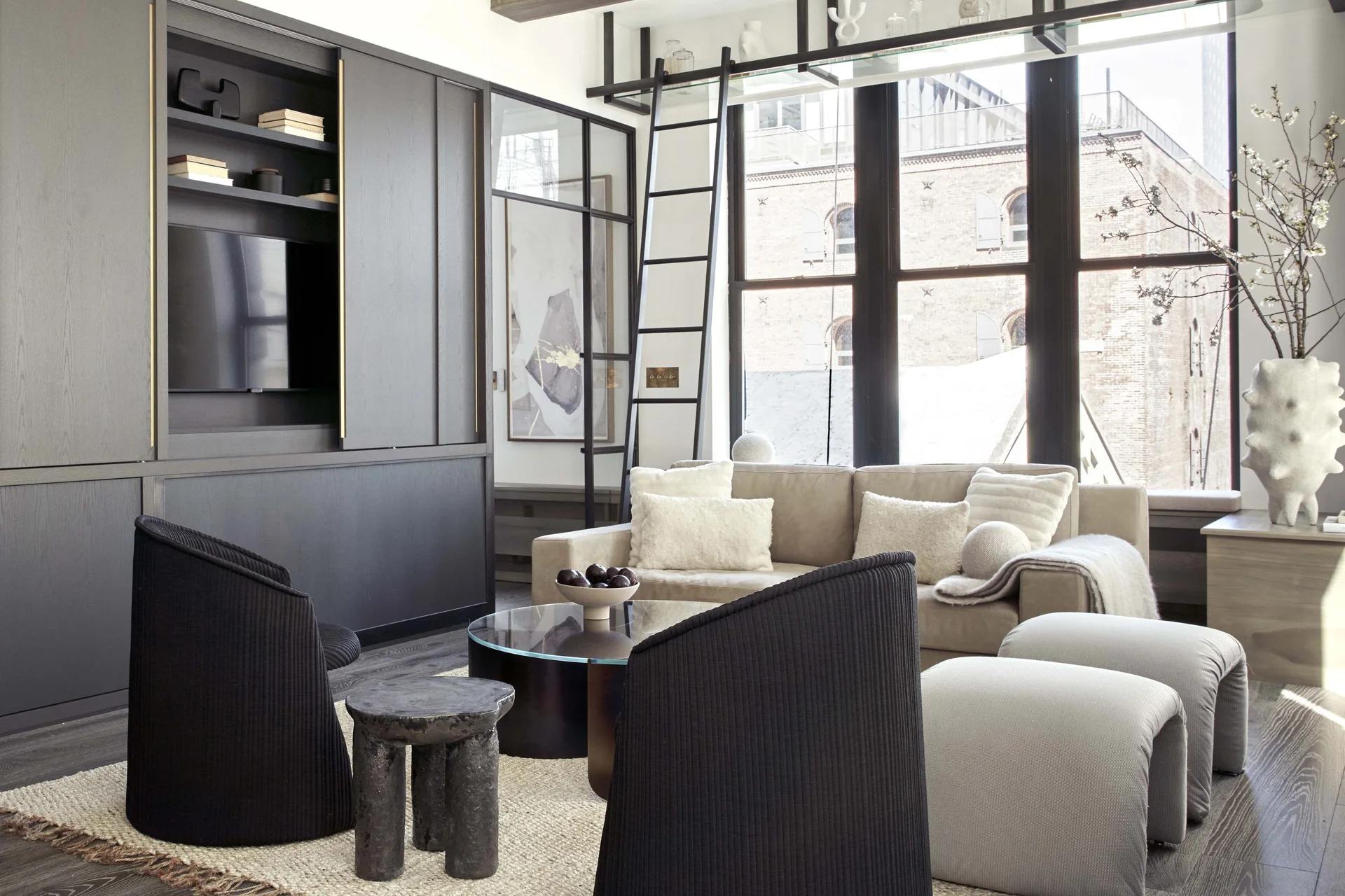 A modern living room features a dark gray built-in wall unit, a beige sofa, ribbed black accent chairs, and a black library ladder by a large window.