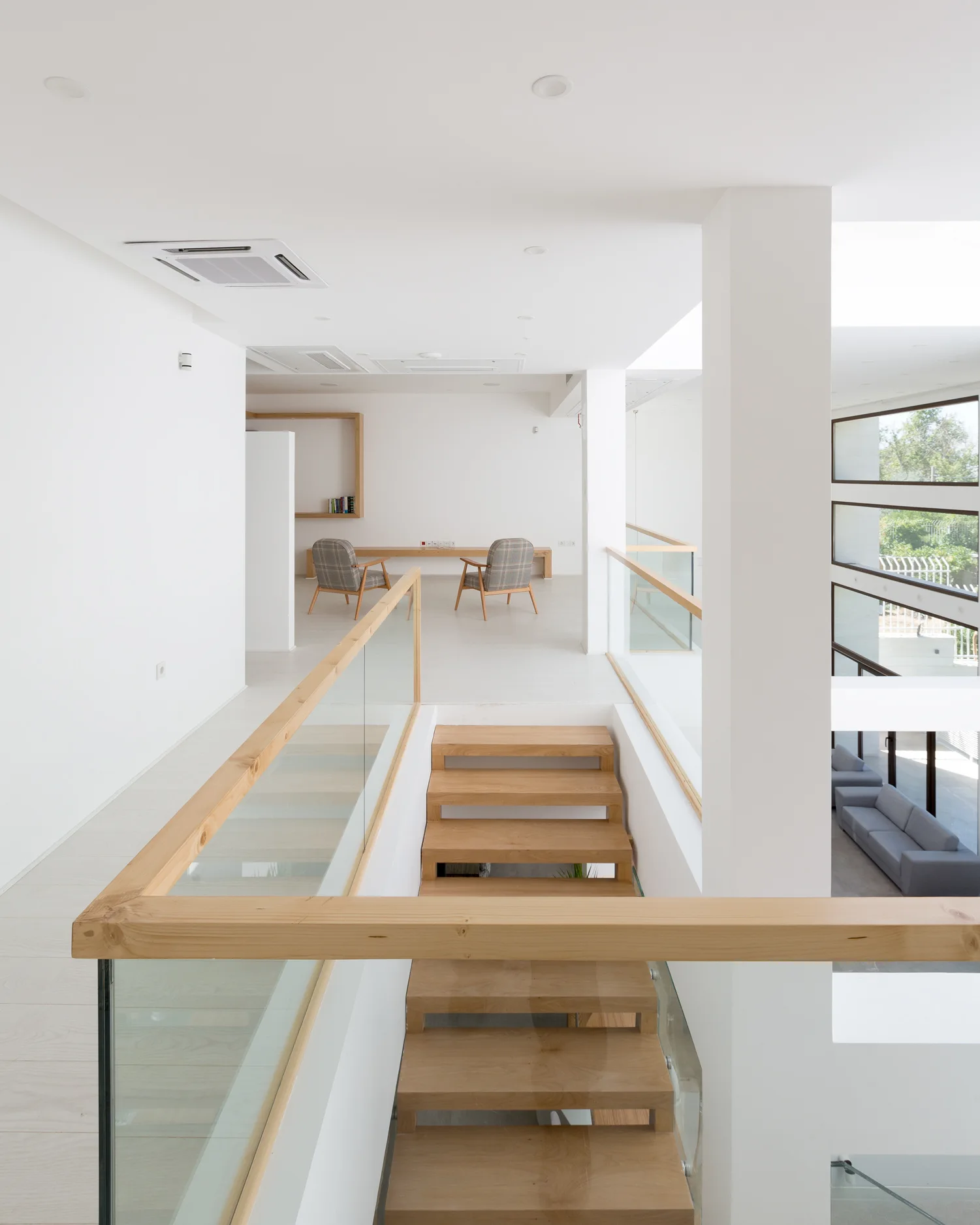A modern wooden staircase with glass railings leads up to a bright white landing with two armchairs and a bench.