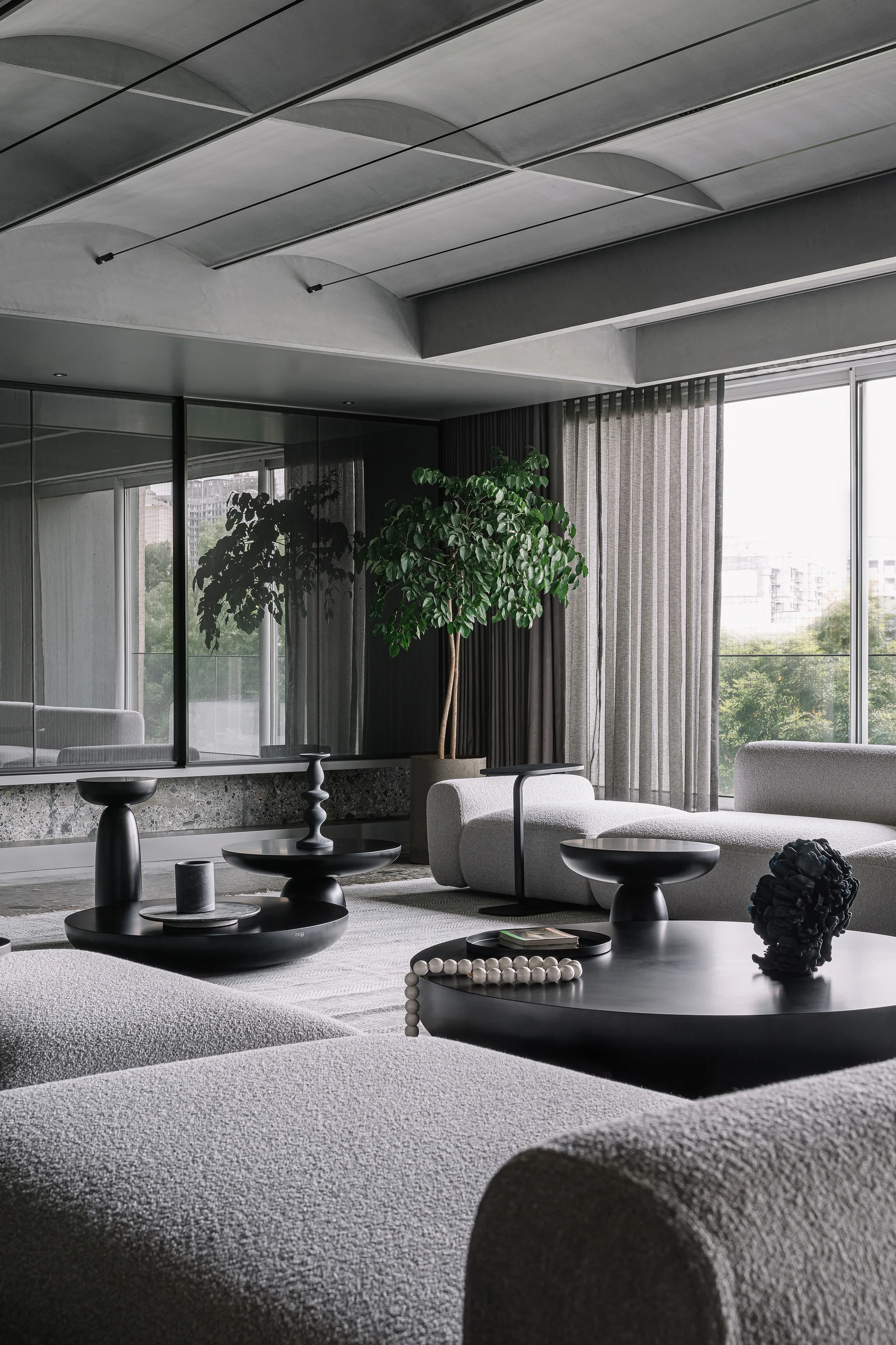 A modern living room features low grey boucle sofas, matte black circular coffee tables, a large potted tree, and a vaulted concrete ceiling.
