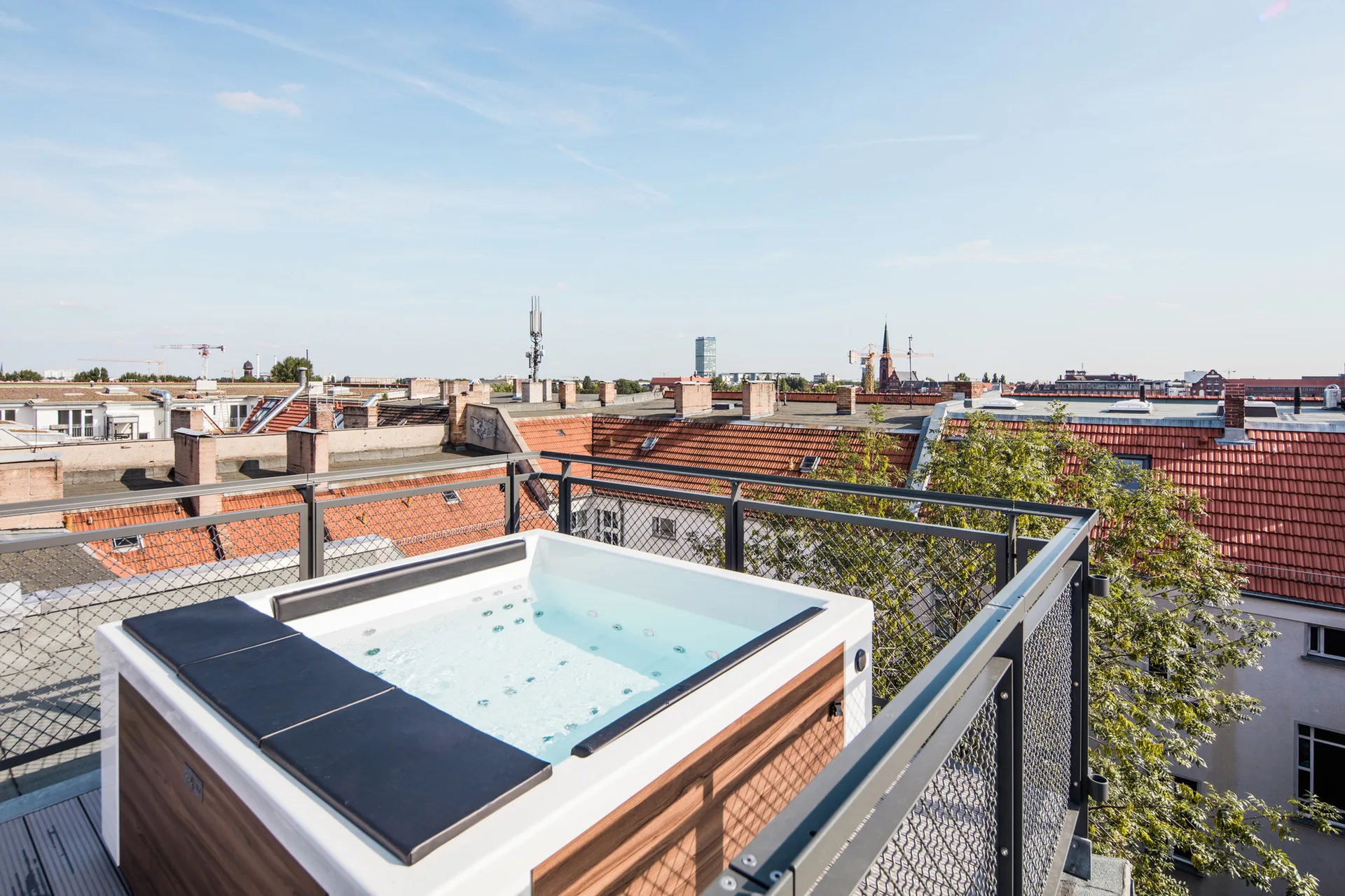A rectangular hot tub with wood-grain side panels sits on a grey-decked rooftop balcony surrounded by a grey metal mesh railing under a blue sky.