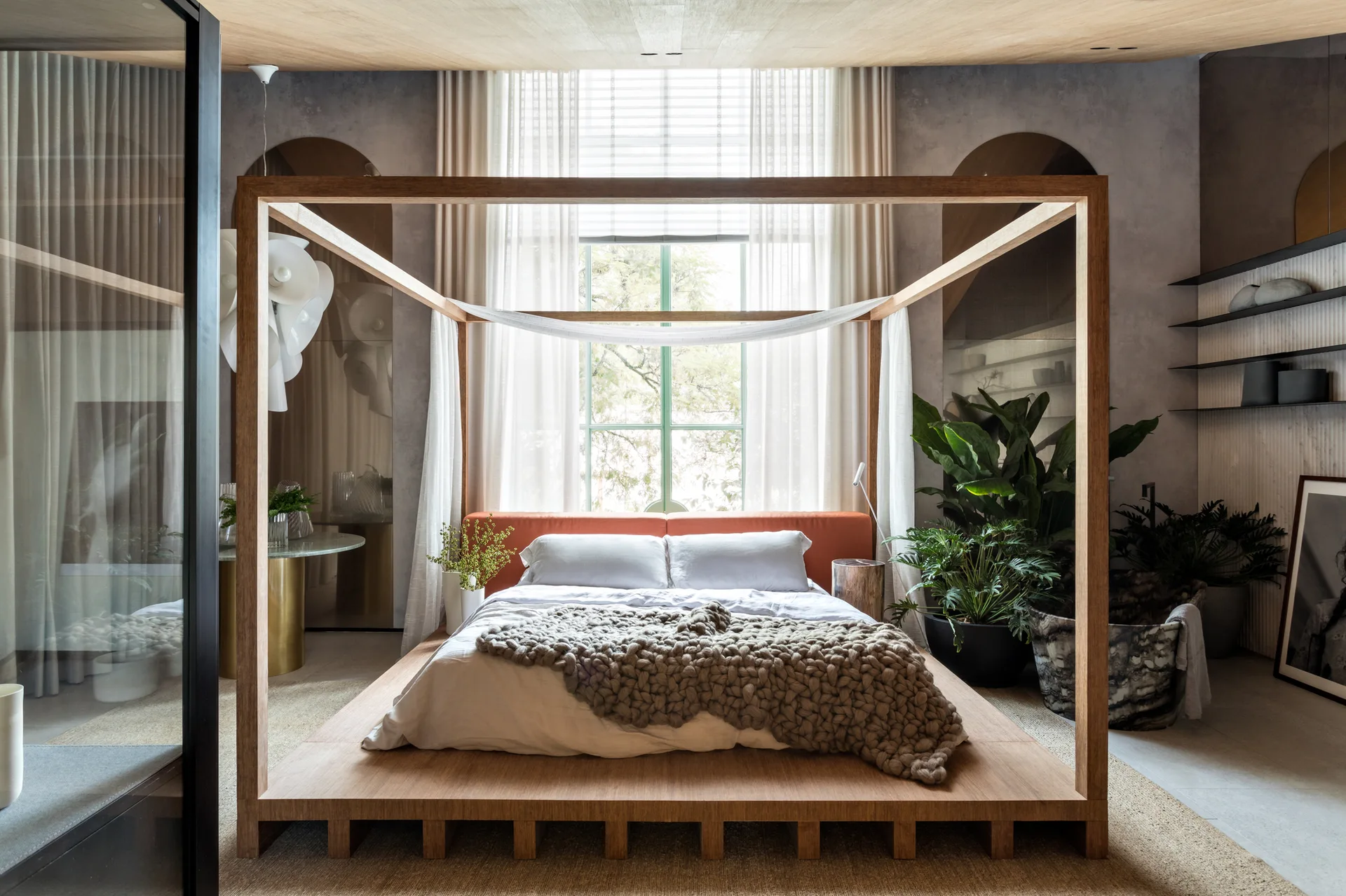 A modern bedroom with a large wooden platform canopy bed, a terracotta headboard, and a chunky knit blanket in front of a window with sheer curtains.