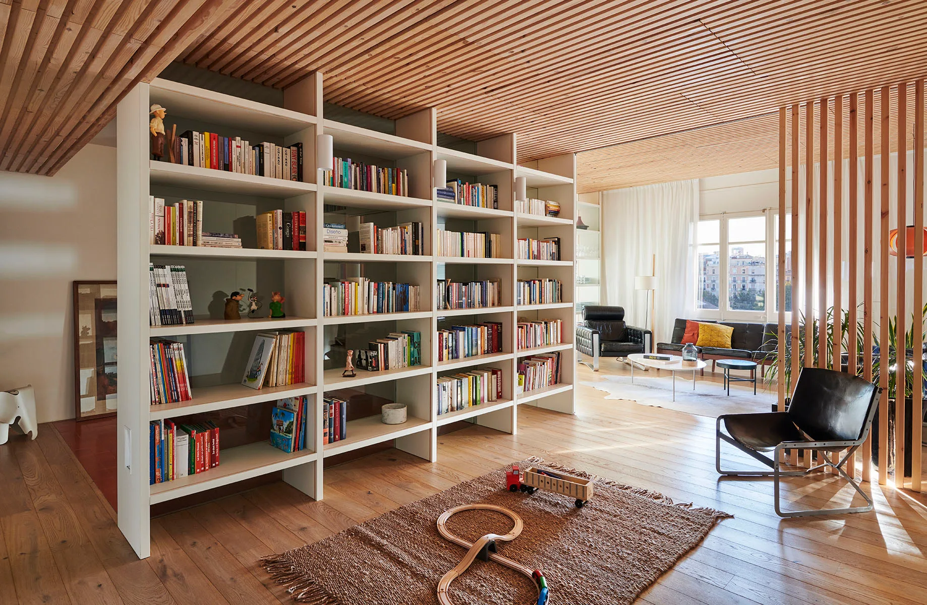 A spacious living room featuring a wood slatted ceiling, a large white bookshelf room divider, wooden floors, and mid-century modern seating.