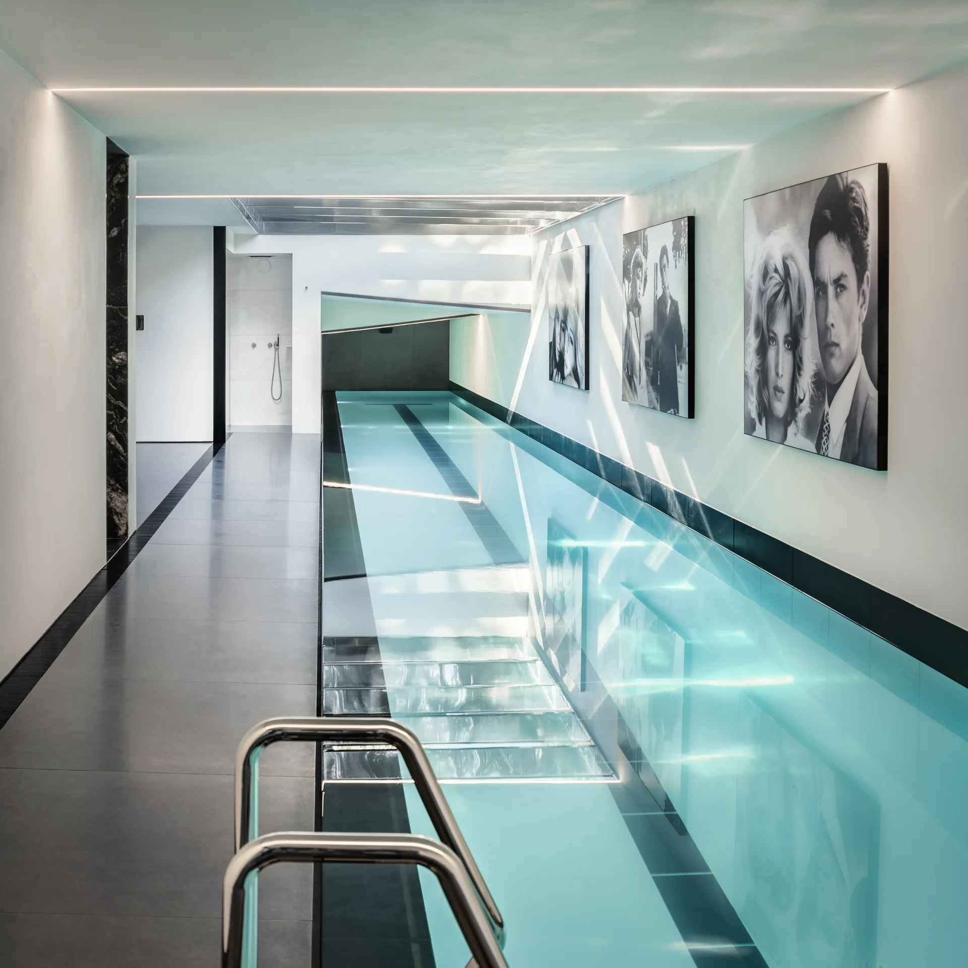 A minimalist indoor lap pool features turquoise water, dark grey tiled flooring, linear ceiling lights, and framed black-and-white portraits on the wall.