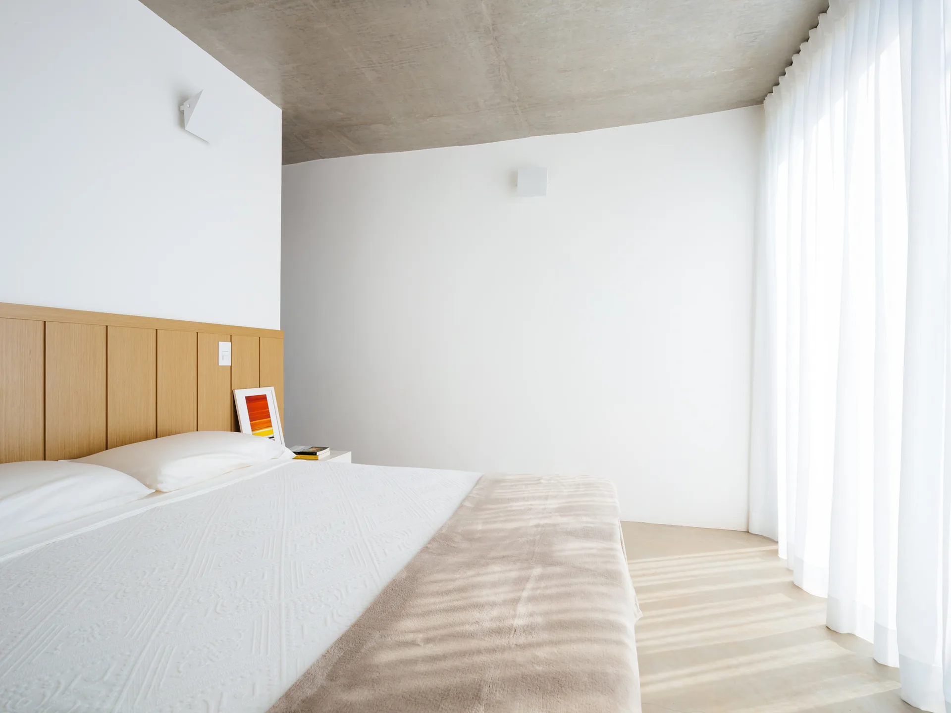 A minimalist bedroom featuring a light wood headboard wall, an exposed concrete ceiling, and white bedding near sheer white curtains.
