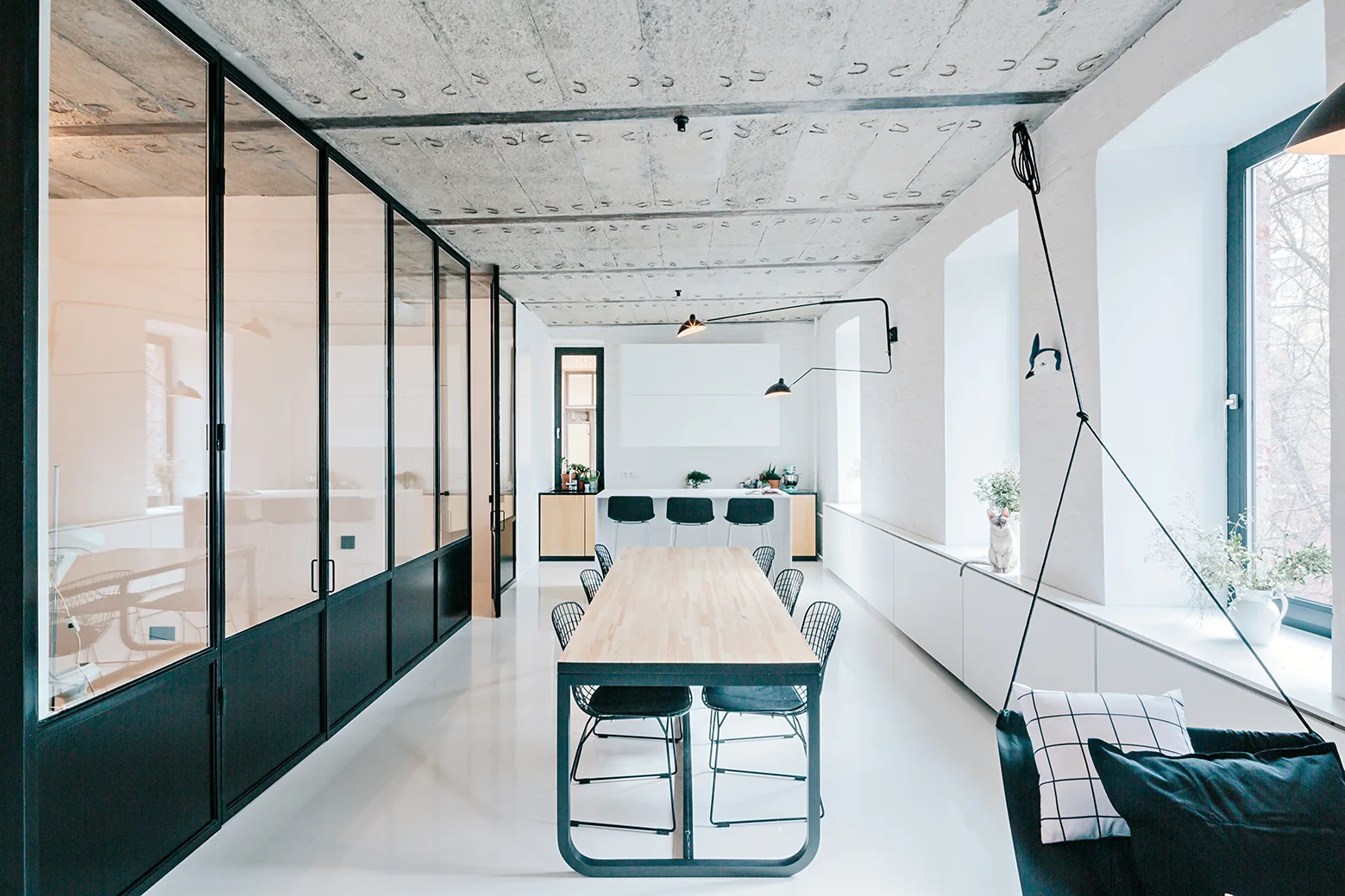 A minimalist dining room with an exposed concrete ceiling, a long wood table, and black-framed glass partition walls.