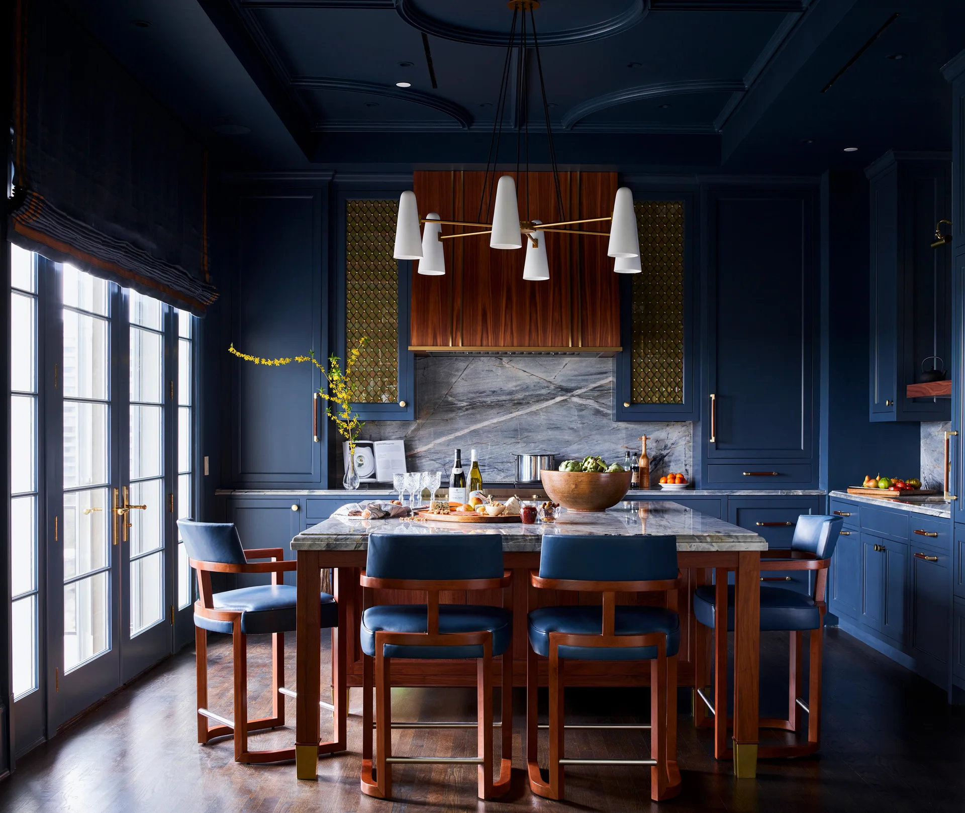 A modern traditional kitchen features deep navy blue cabinetry, a coffered ceiling, a stone-topped island with leather bar stools, and a wood range hood.