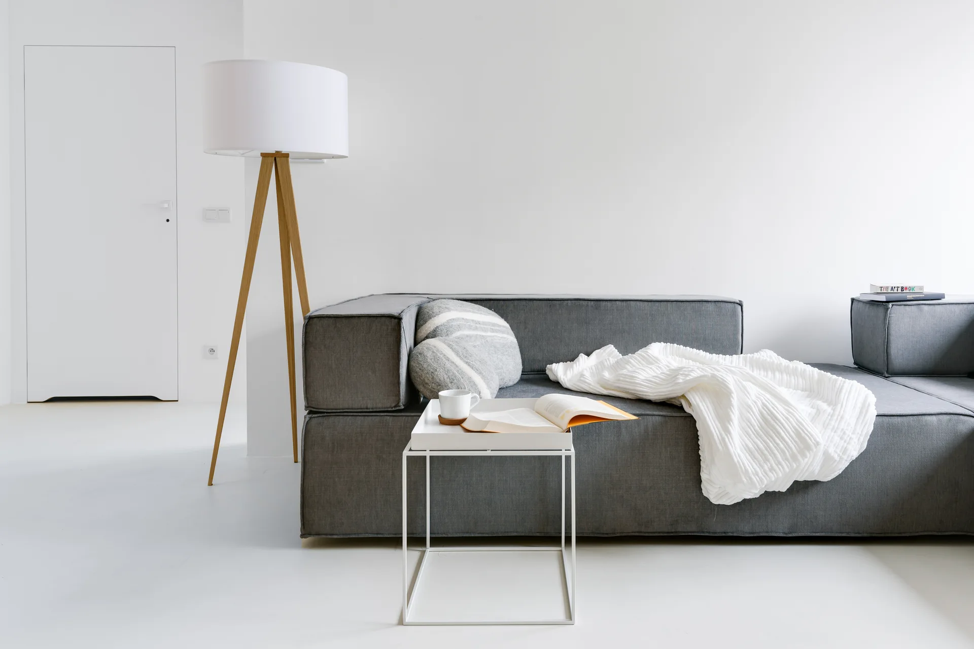 A low-profile grey sectional sofa sits against a white wall next to a wooden tripod floor lamp and a white metal tray table.