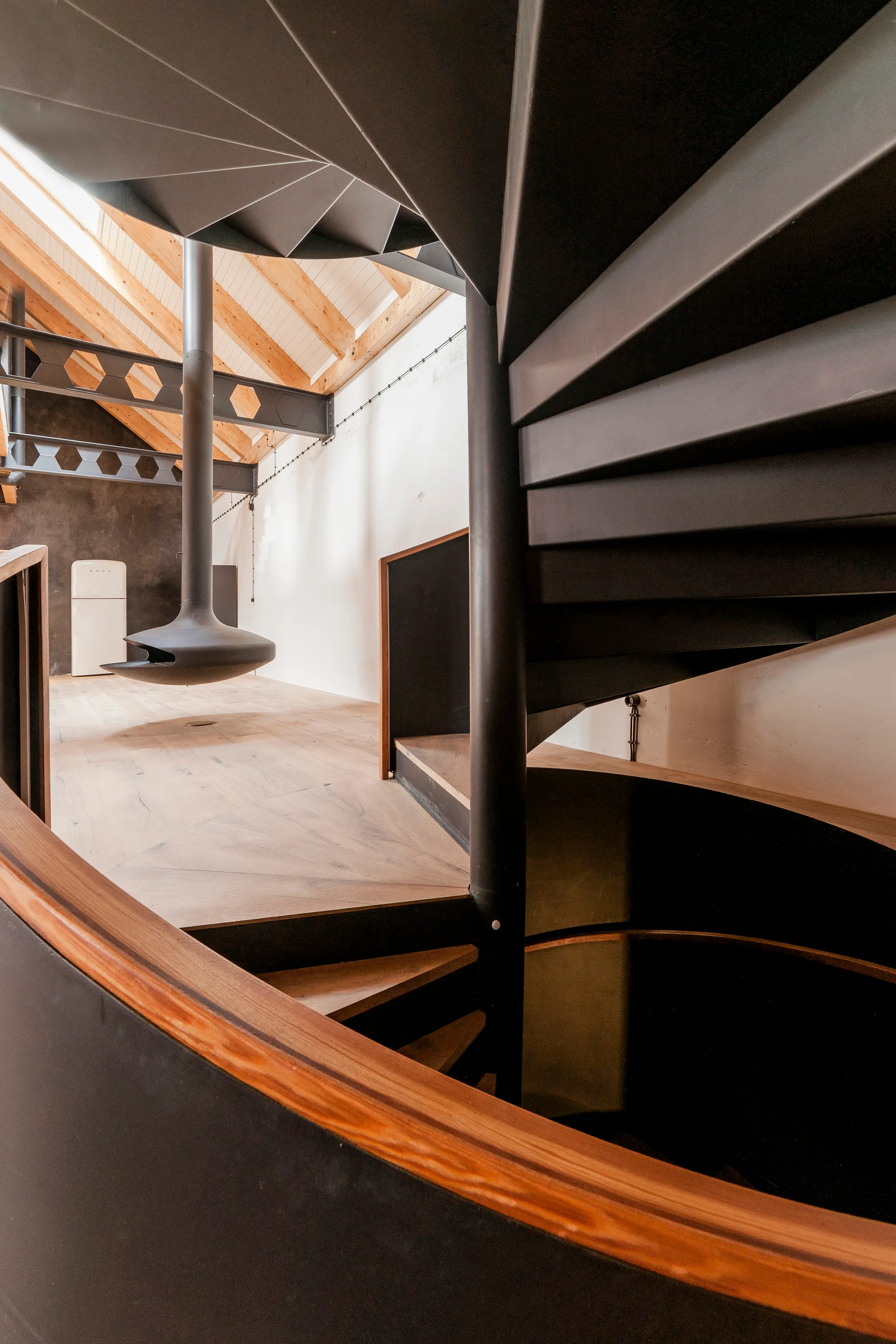 A black metal spiral staircase winds through an industrial loft featuring a suspended fireplace and exposed wooden ceiling beams.