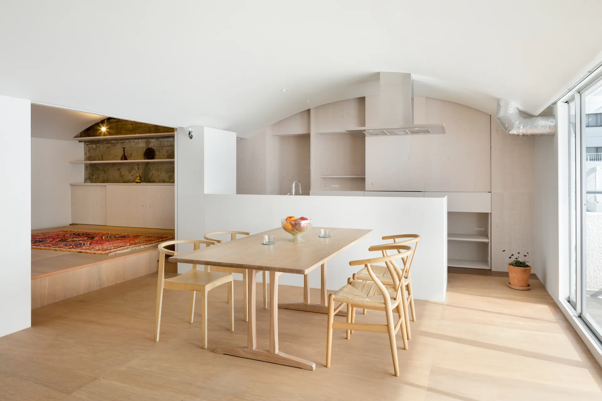 A bright dining area features a light wood table, woven-seat chairs, and a curved white ceiling next to a minimalist kitchen with plywood cabinetry.