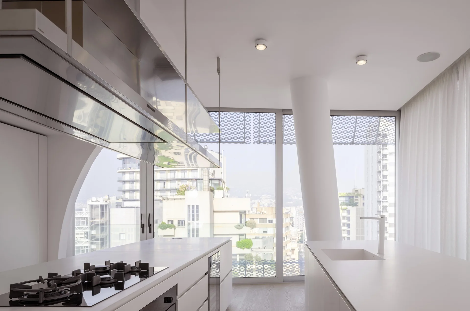 A minimalist white kitchen features parallel islands, a large polished stainless steel range hood, and floor-to-ceiling windows overlooking a city.