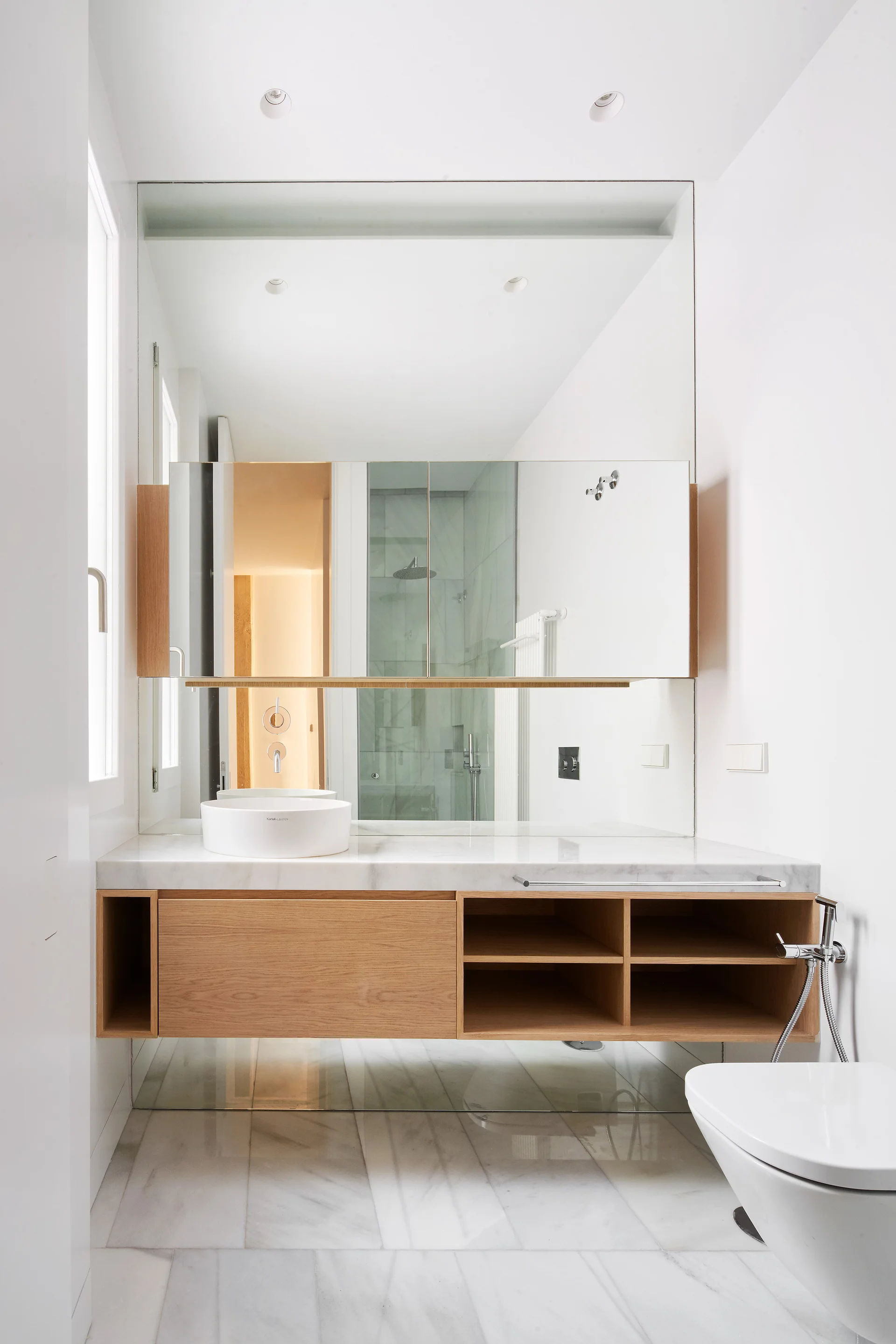 A minimalist bathroom features a floating wooden vanity with a white marble countertop, round vessel sink, and a large wall-mounted mirror.