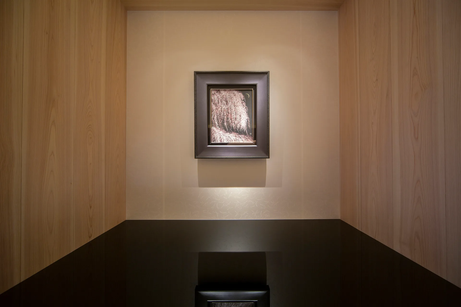 A framed painting hangs centered inside a recessed wall alcove lined with light wood paneling above a glossy black reflective countertop.