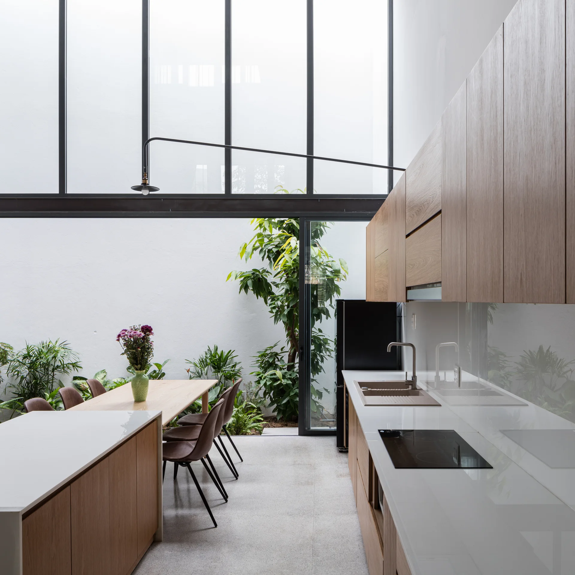 A modern kitchen with wood cabinets and a white island next to a dining table facing a double-height glass wall looking into a courtyard garden.