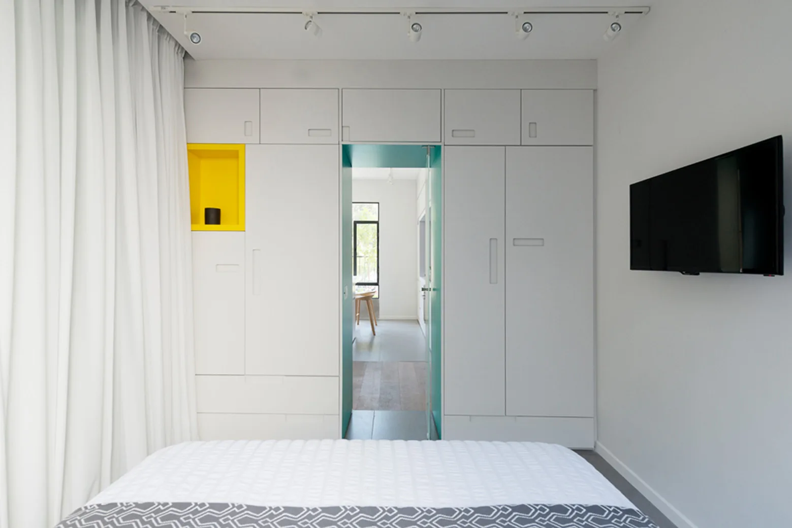 A minimalist white bedroom featuring a floor-to-ceiling built-in wardrobe with a yellow display niche and a doorway framed in teal.
